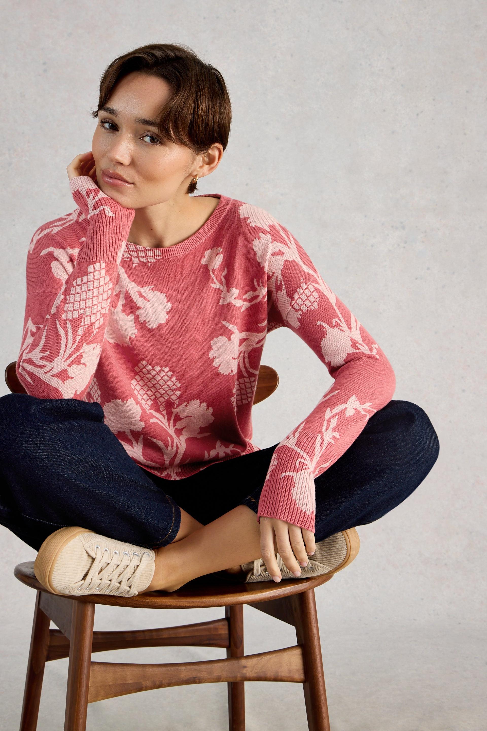 Woman wearing a pink floral sweater sitting on a wooden stool against a plain background