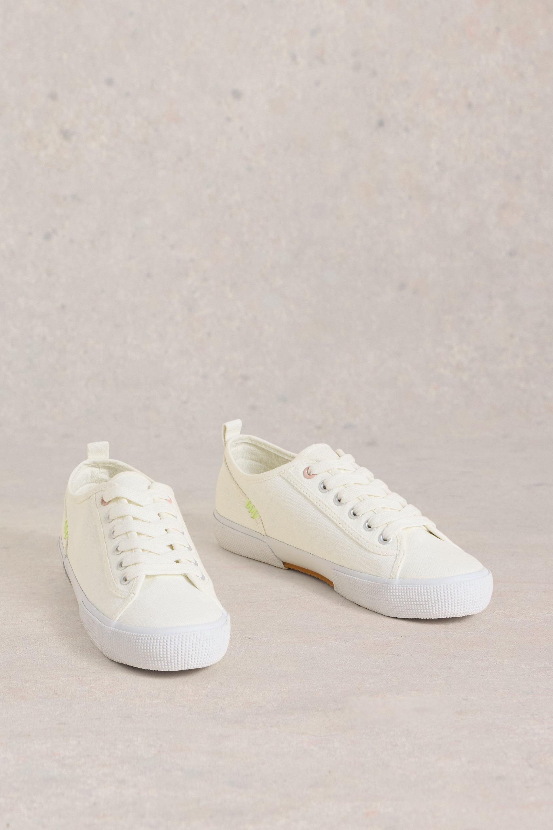Side view of White Stuff Pippa White Canvas Lace Up Trainers in a neutral setting.