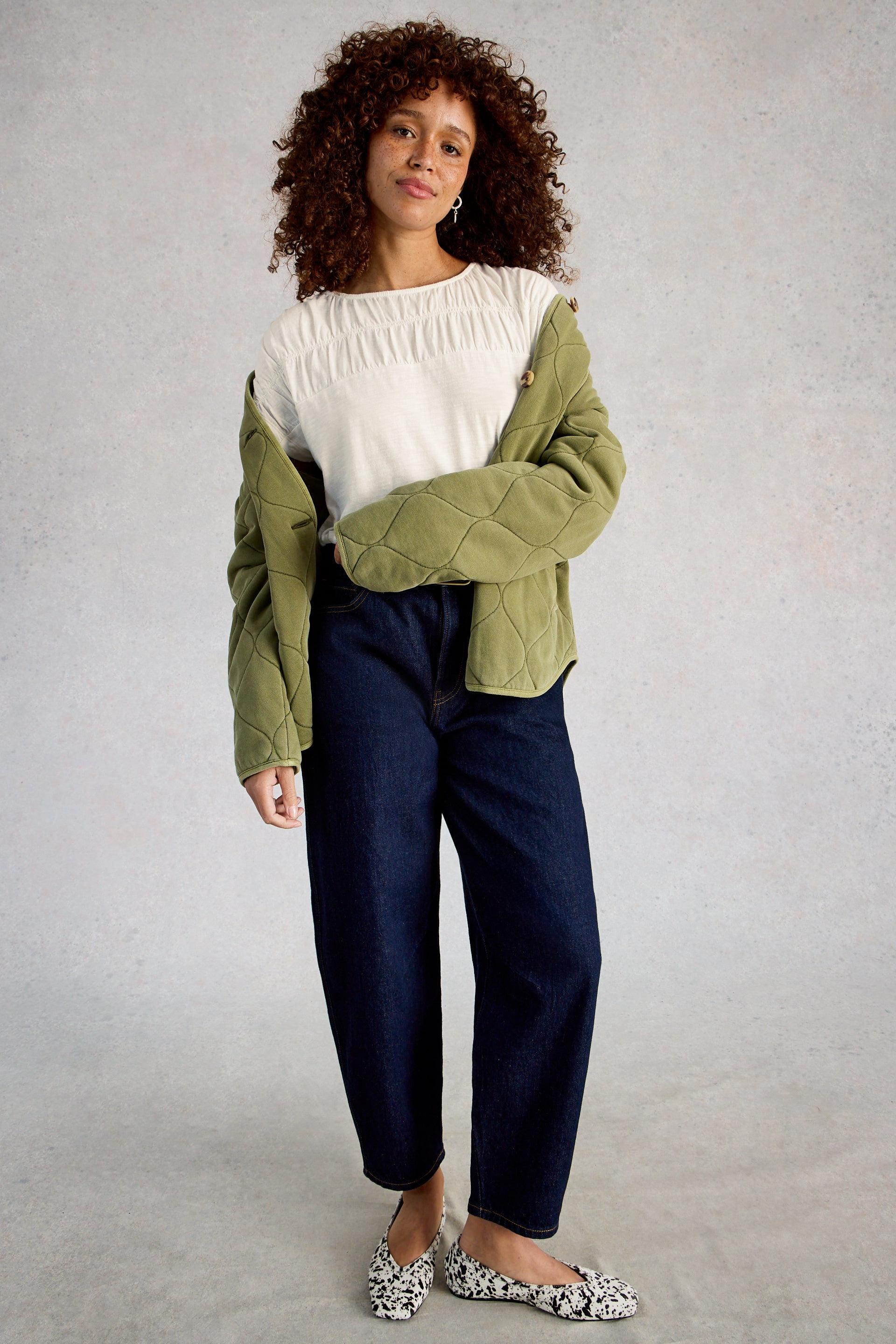 Woman wearing a green quilted jacket over a white organic cotton top with navy pants on a gray background