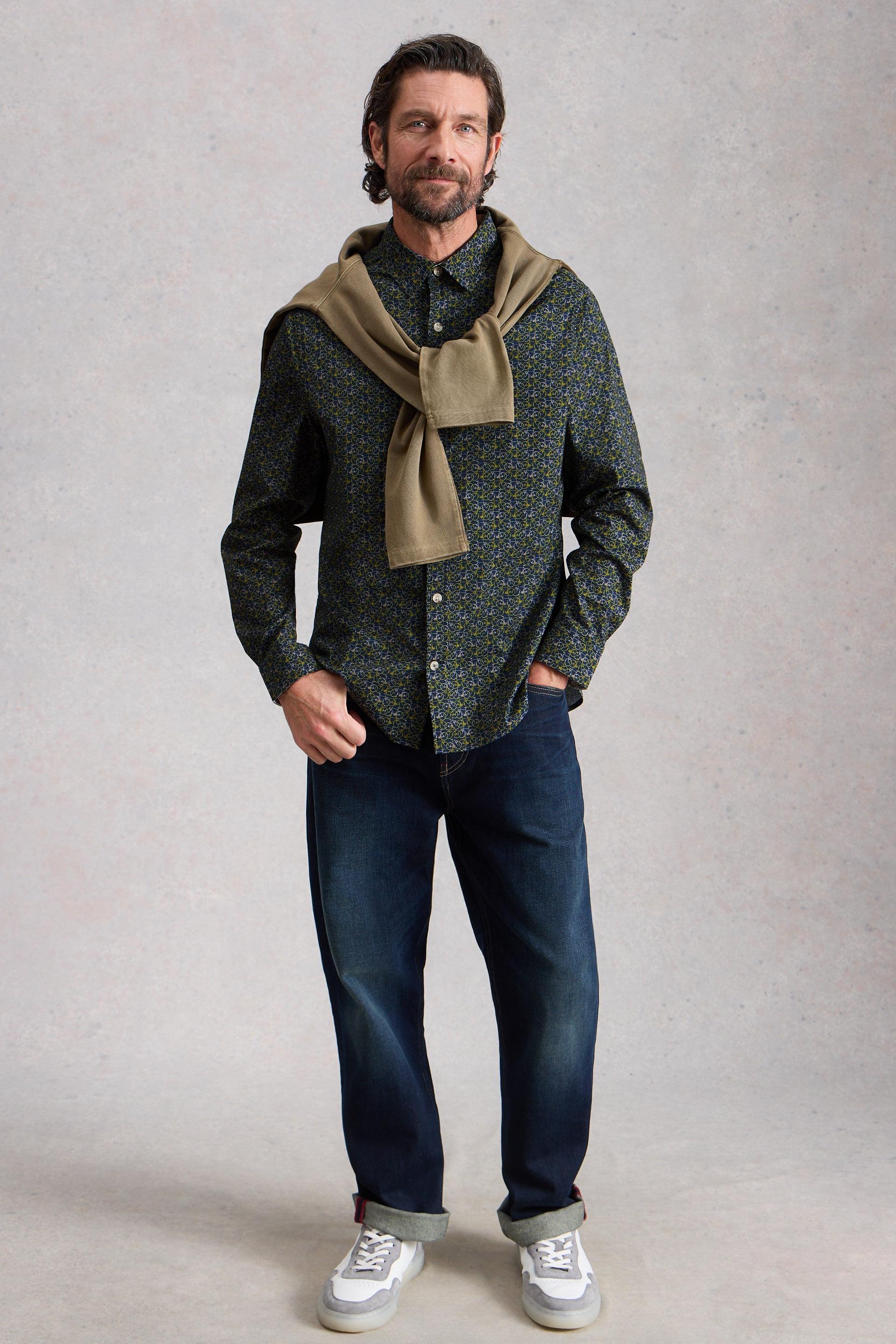 Man wearing a green patterned shirt, blue jeans, and a brown scarf against a plain background