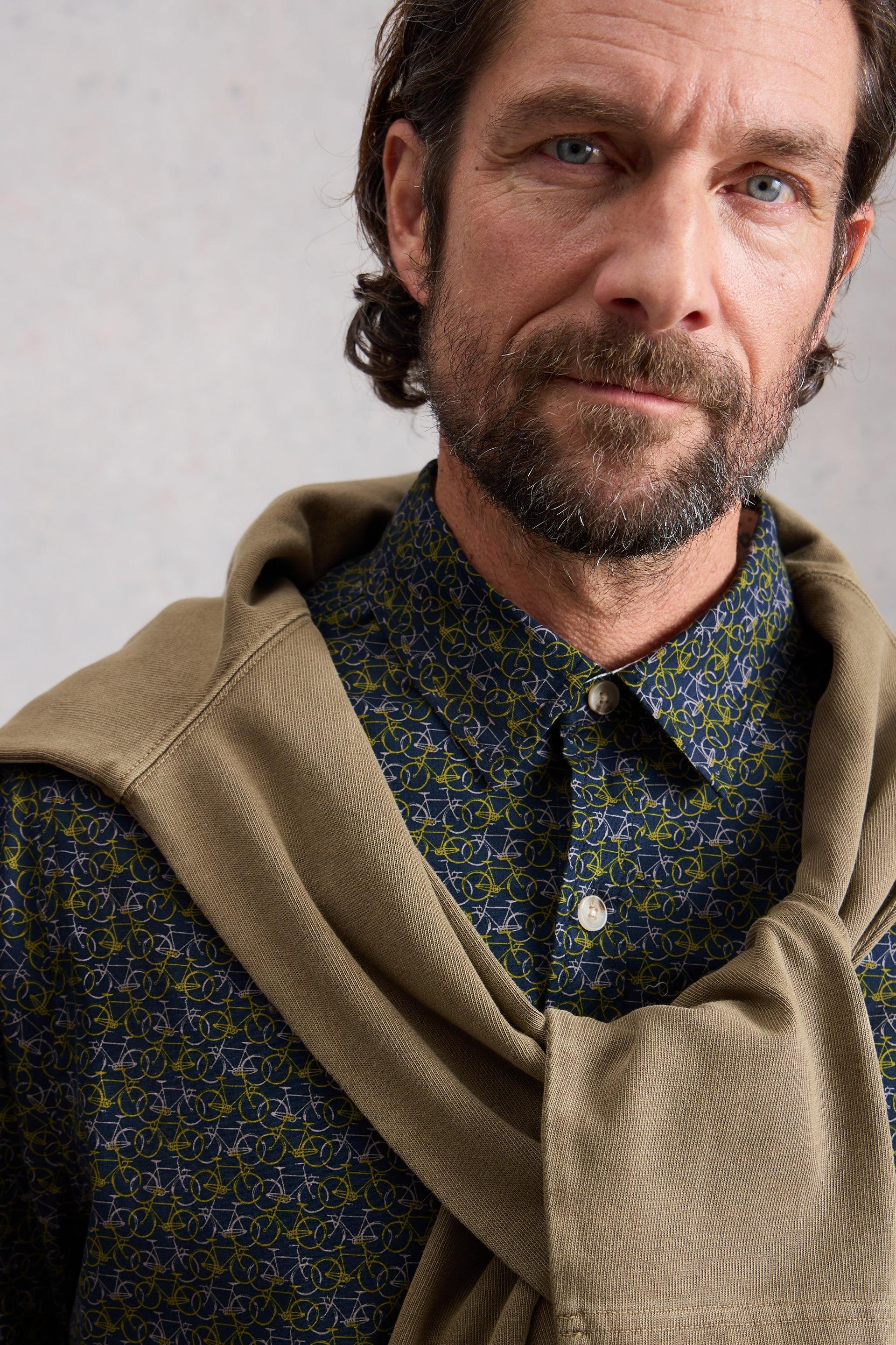 Man wearing a patterned shirt with a beige sweater against a neutral background