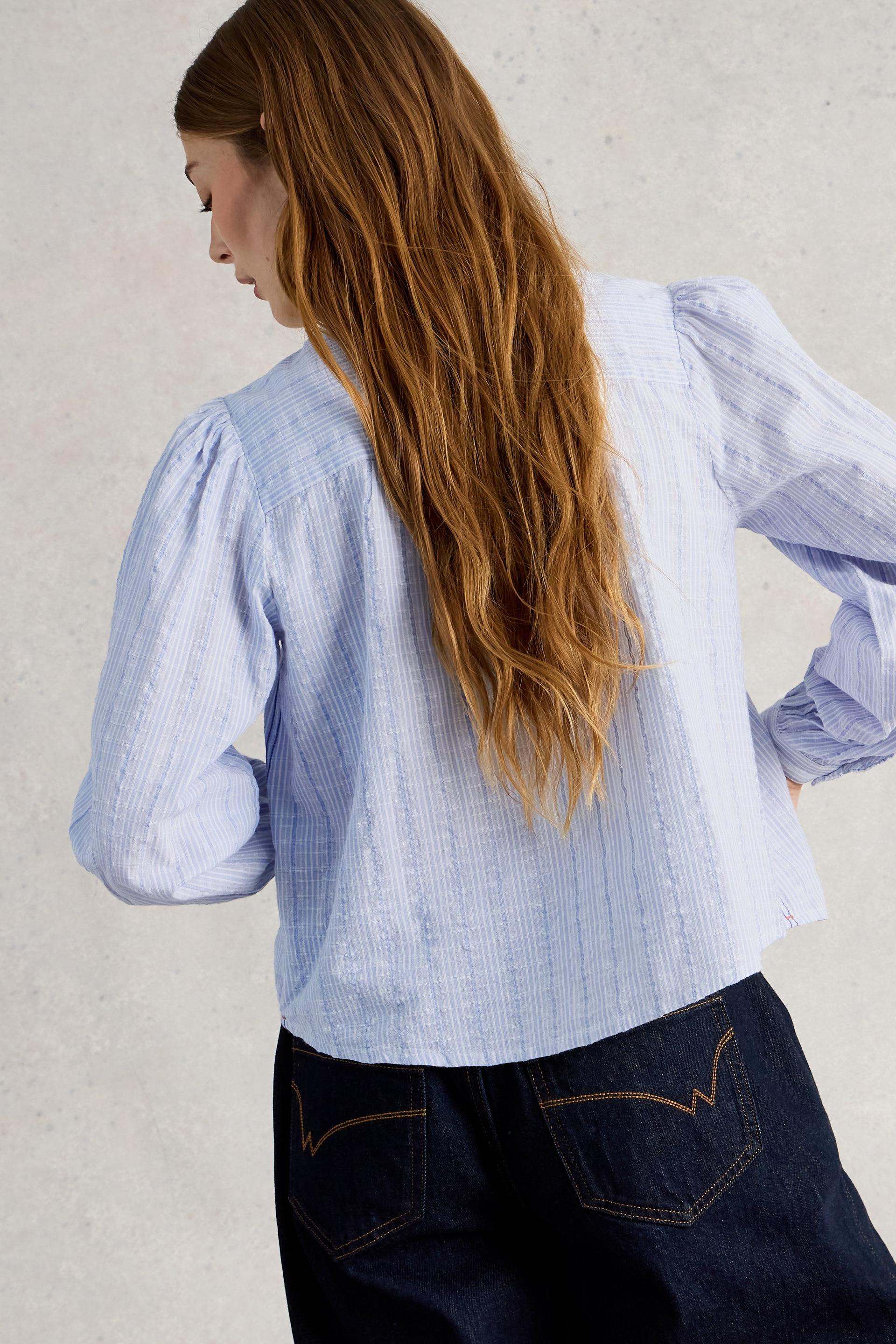 Woman wearing a light blue organic cotton blouse and dark jeans against a neutral background