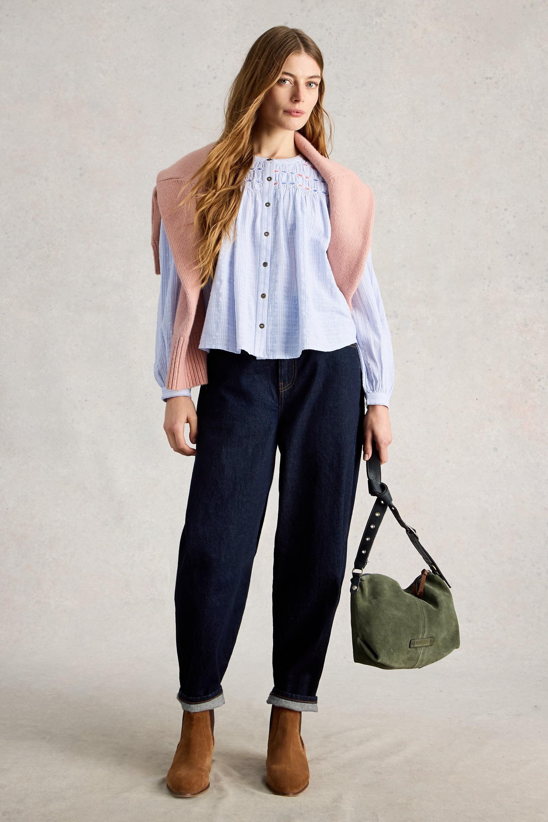 Woman wearing a light blue blouse, dark jeans, and brown boots, holding a green bag against a neutral background.
