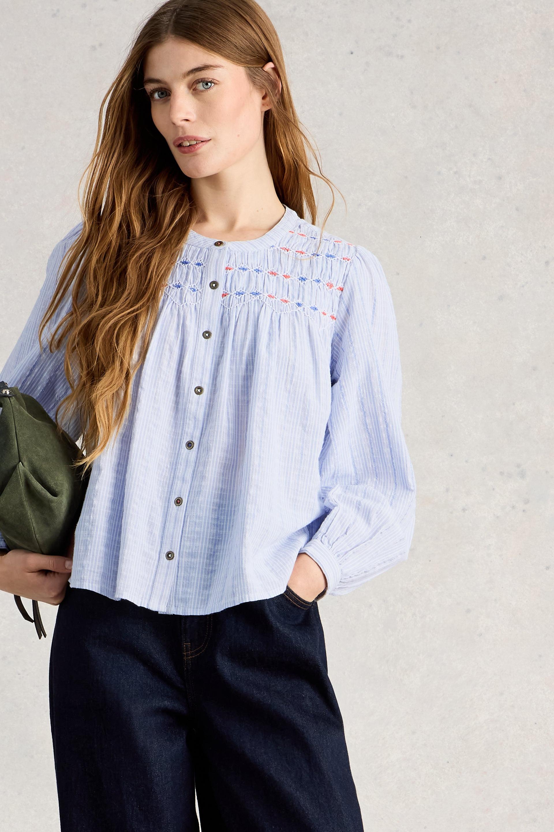 Woman wearing a light blue smock blouse with embroidered details on a neutral background