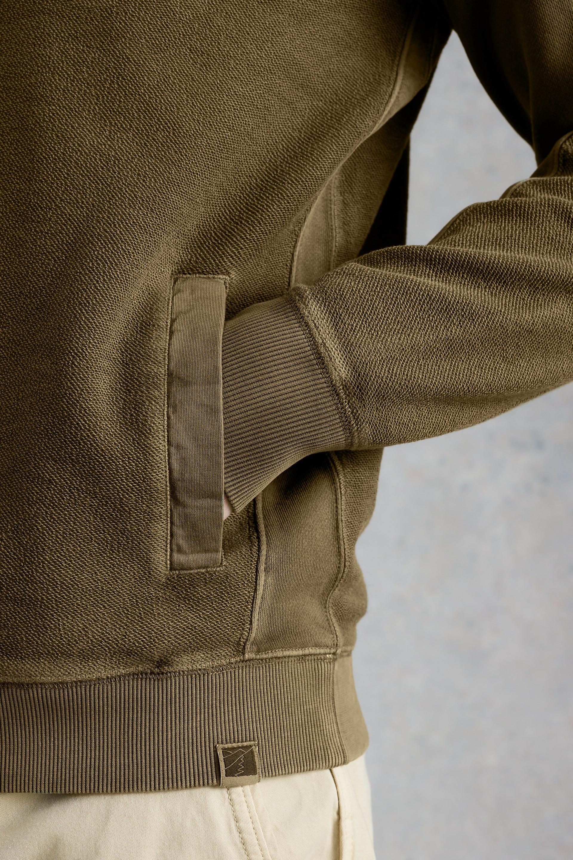 Close-up of a person wearing a green jumper with a visible brand logo on a neutral background