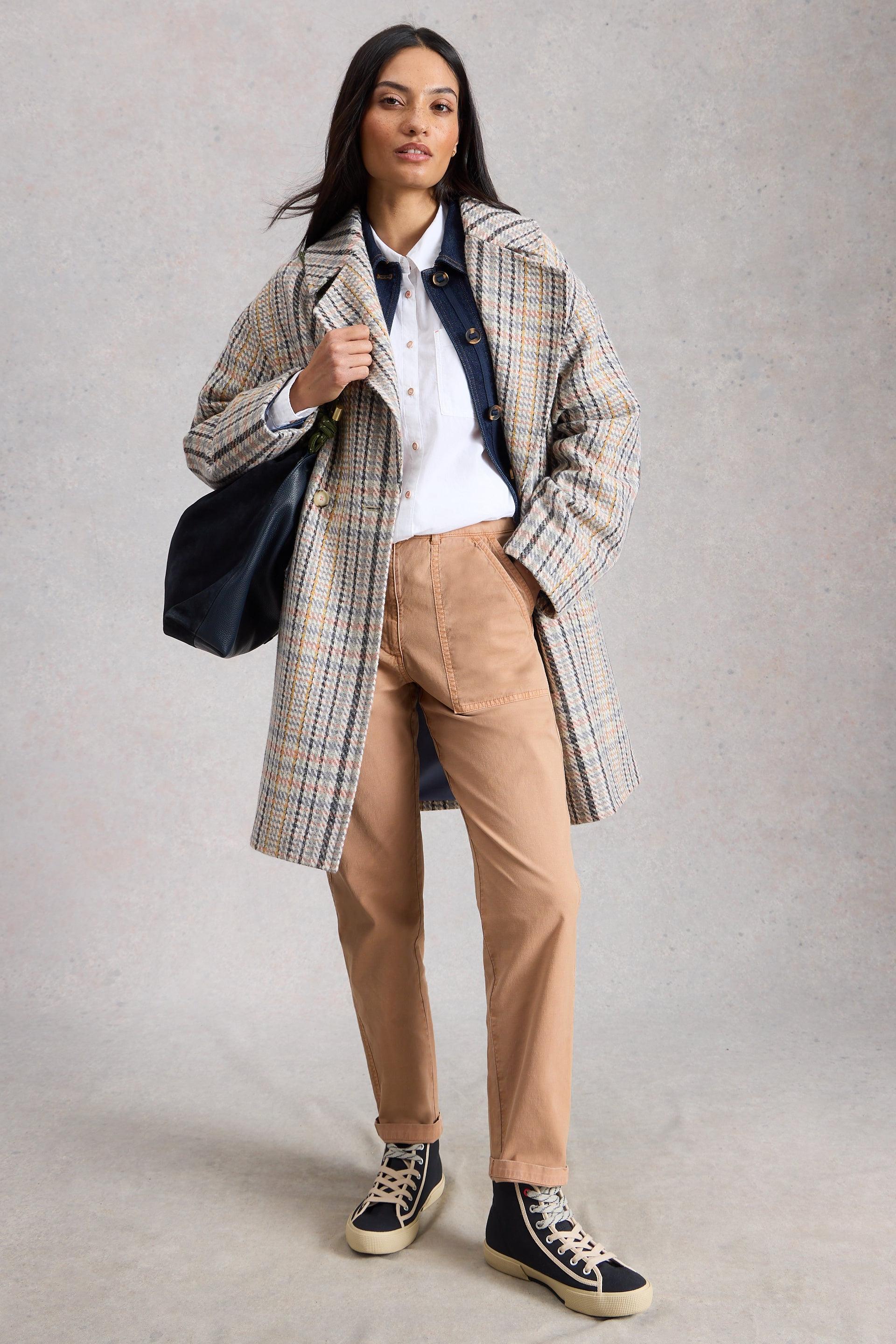 Woman wearing a plaid coat, white shirt, beige organic cotton pants, and black sneakers on a plain background