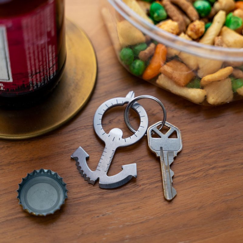 Stainless steel anchor multi tool keychain with a house key on a wooden table near a bottle cap.