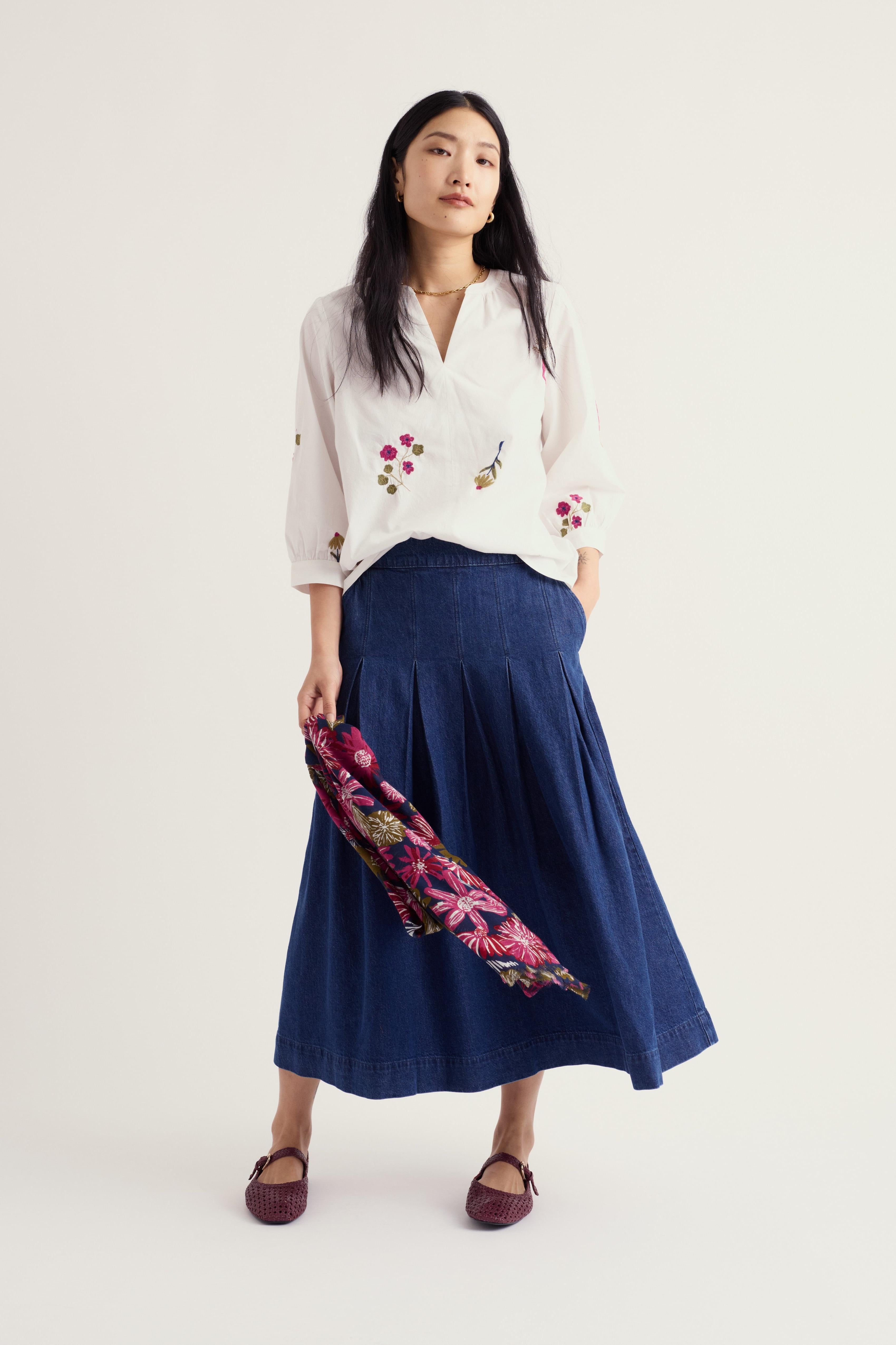 A woman stands against a plain light background wearing a white embroidered blouse, a long navy skirt, and dark burgundy flats, while holding a pink floral scarf.