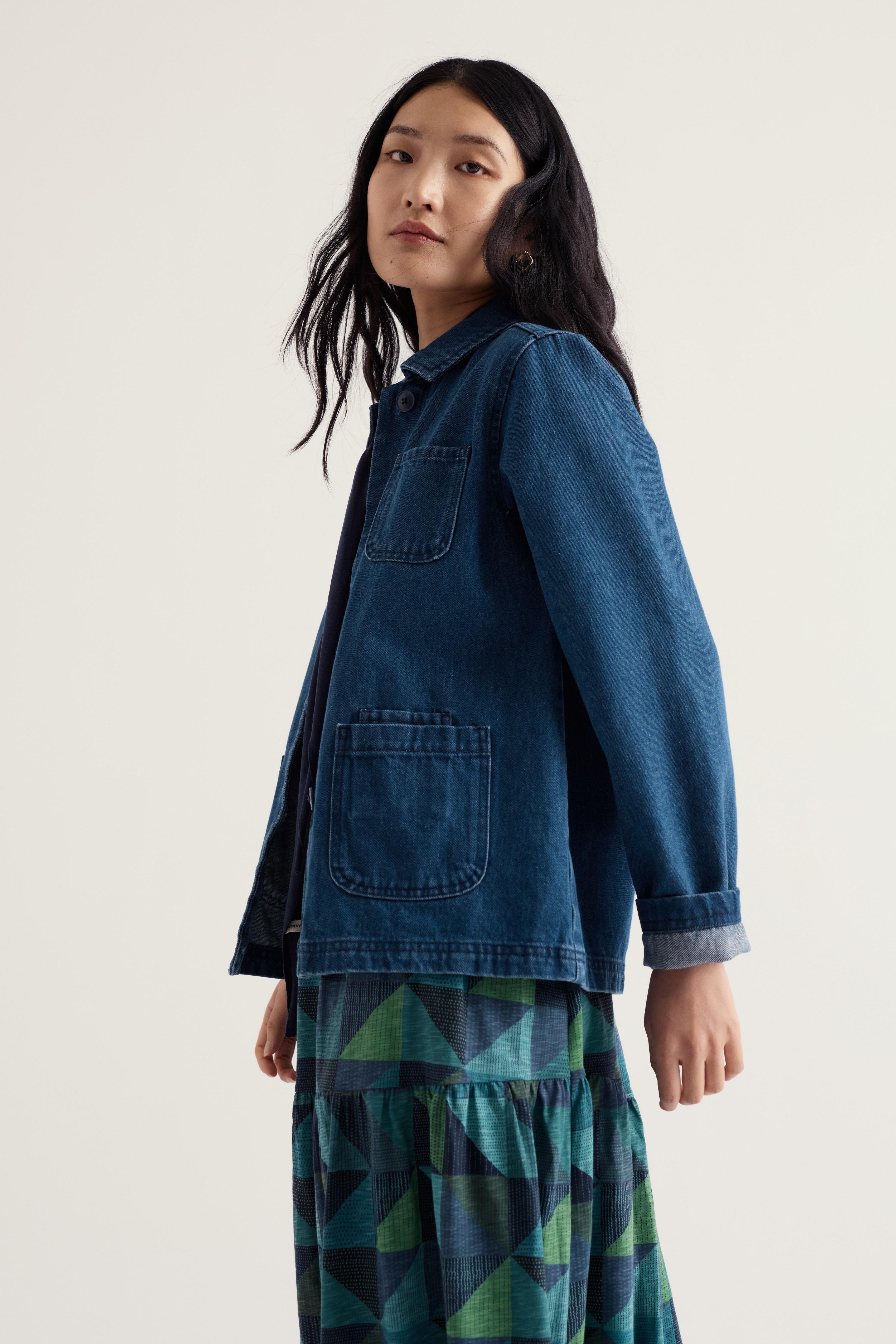 A model wearing a dark blue denim utility jacket with front pockets, layered over a green and blue geometric patterned skirt, photographed against a plain light gray studio background.