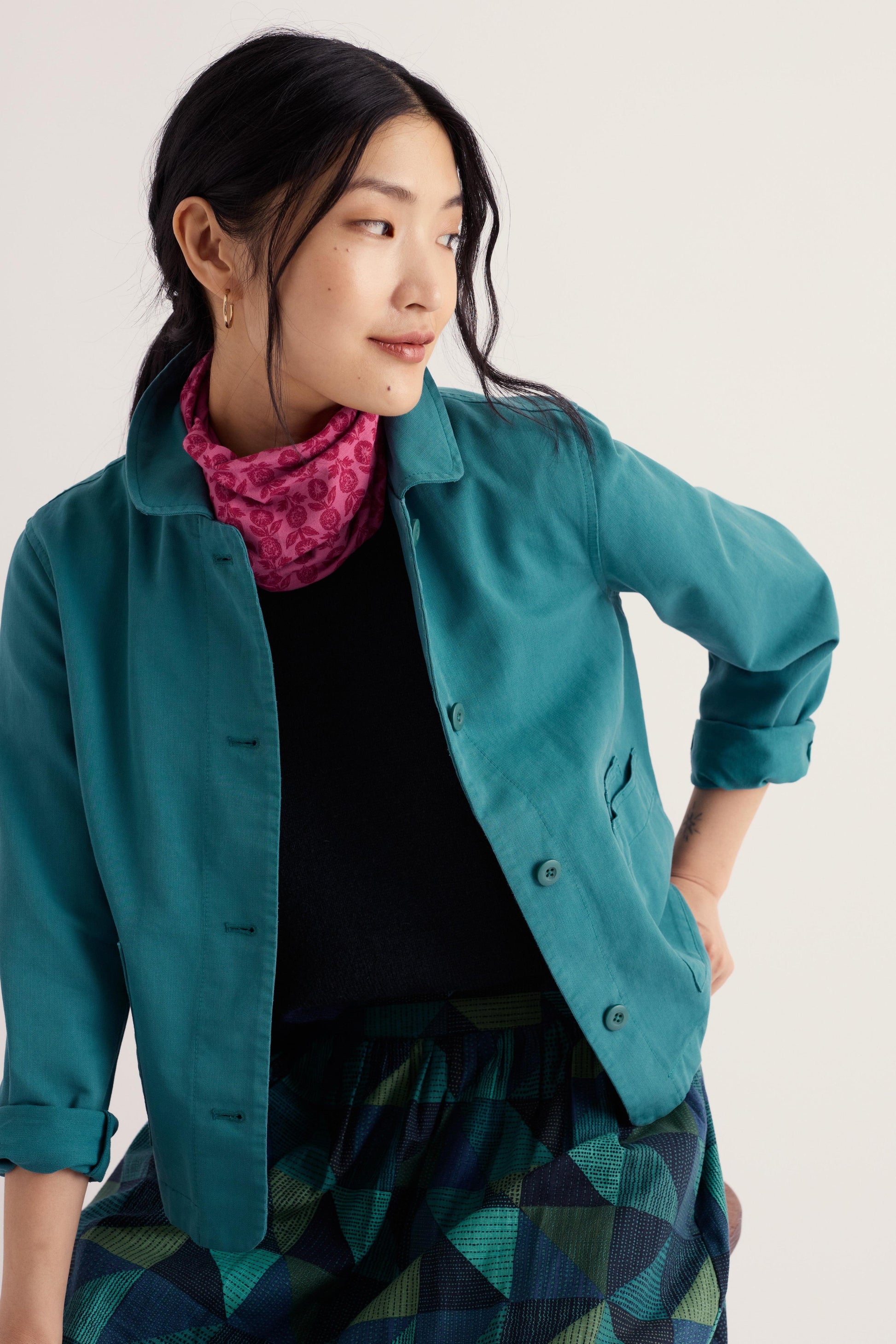 A woman wearing a teal button-up shirt jacket over a black top and a pink patterned scarf, paired with a green and black geometric print skirt, against a plain light background.