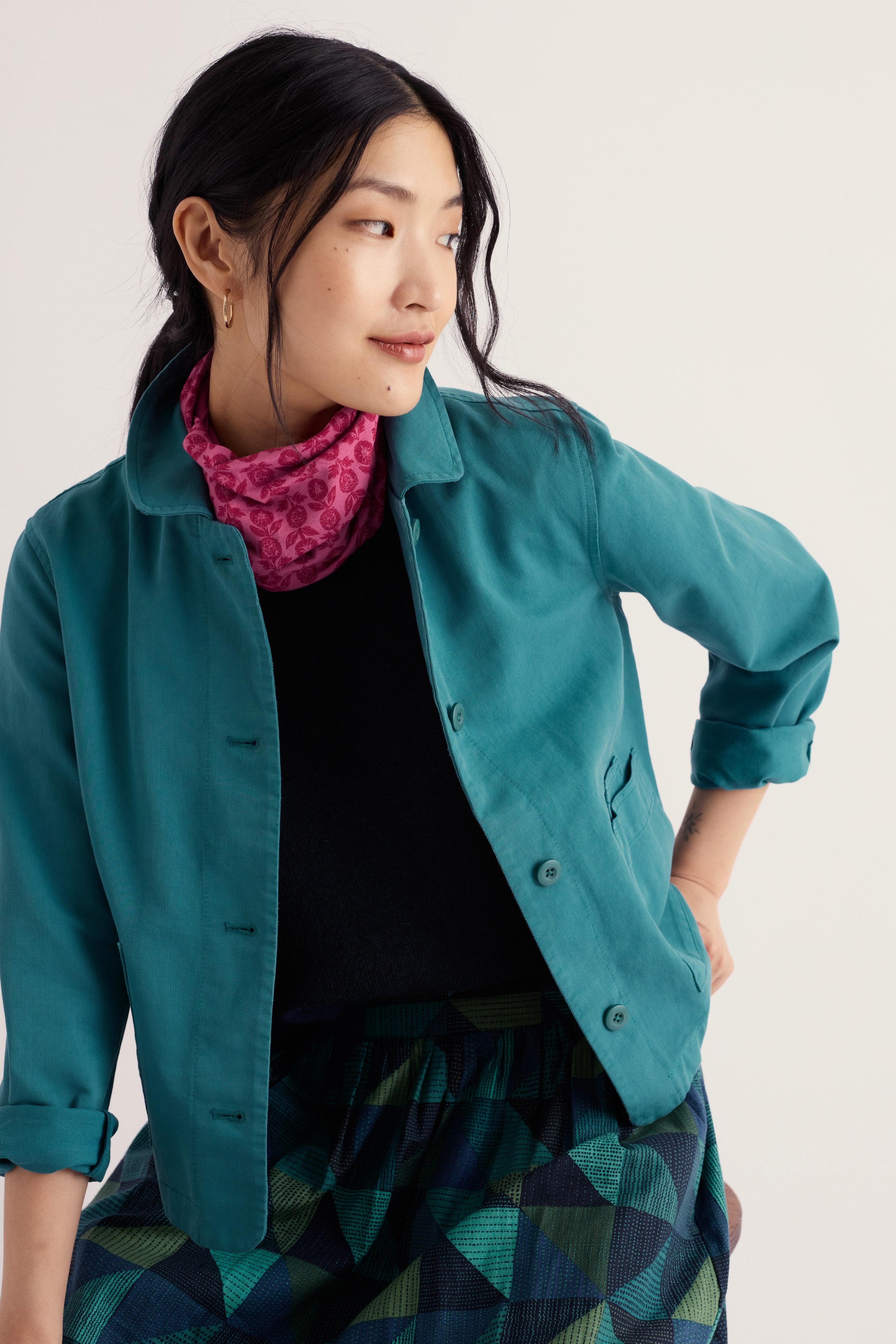 A woman wearing a teal button-up shirt jacket over a black top and a pink patterned scarf, paired with a green and black geometric print skirt, against a plain light background.