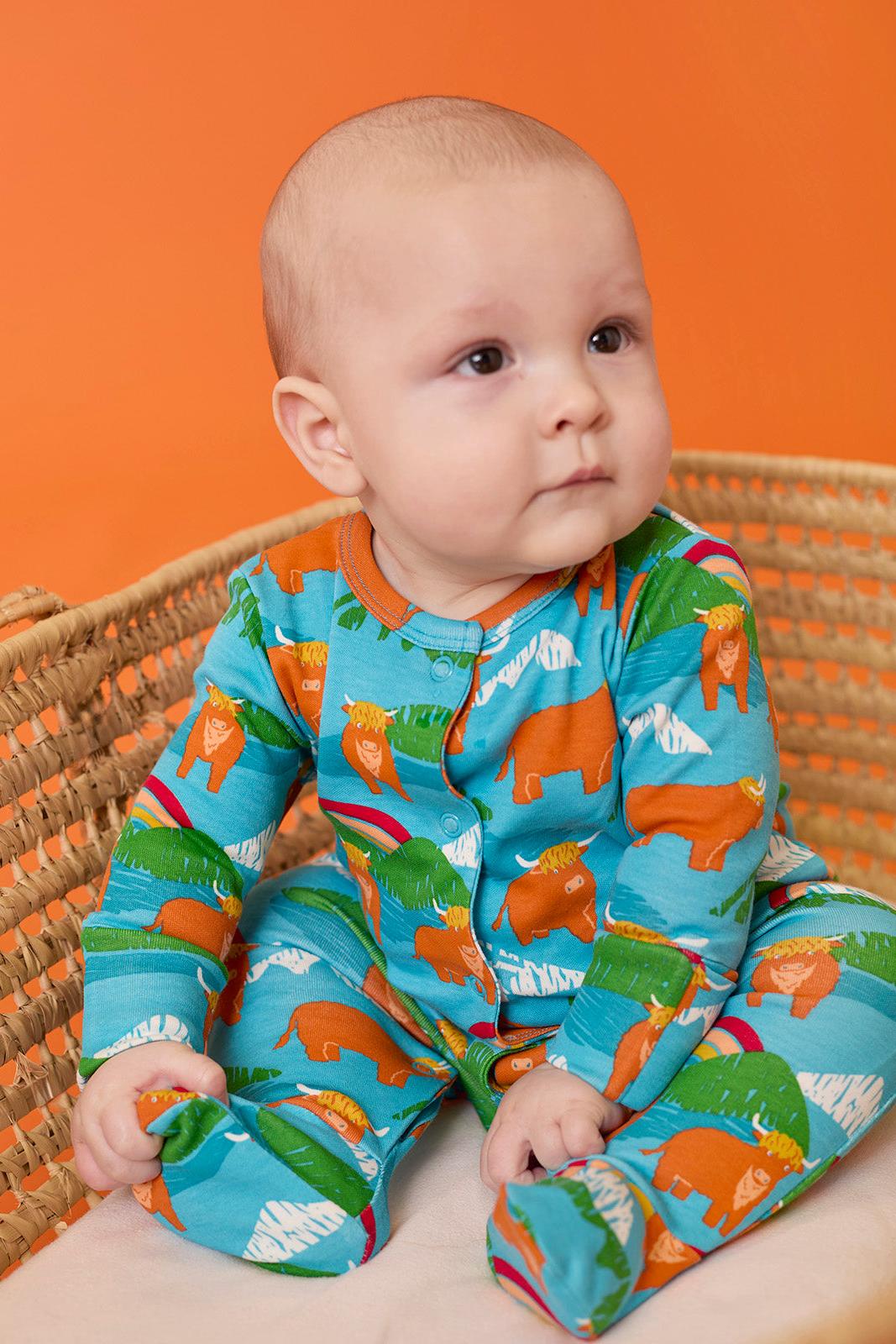 A cute baby boy wearing a blue organic cotton babygro with a farm cow print sitting in a wicker basket.