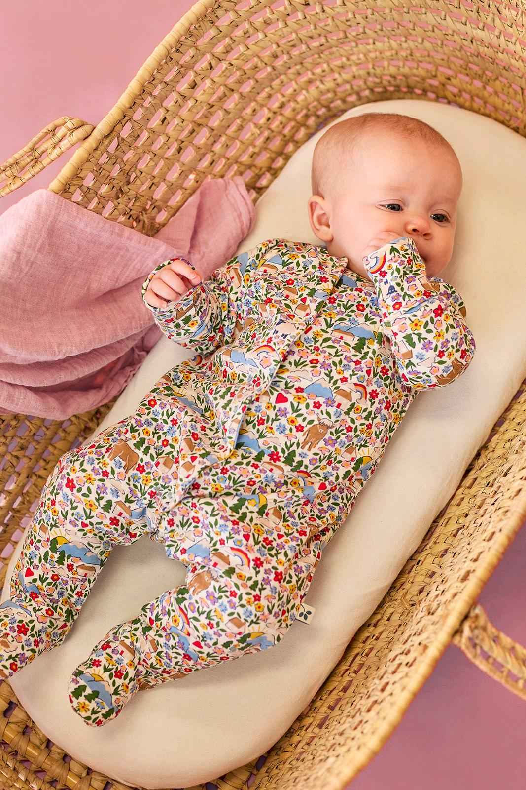 A baby girl wearing a floral Frugi organic cotton babygro lying in a woven wicker moses basket.