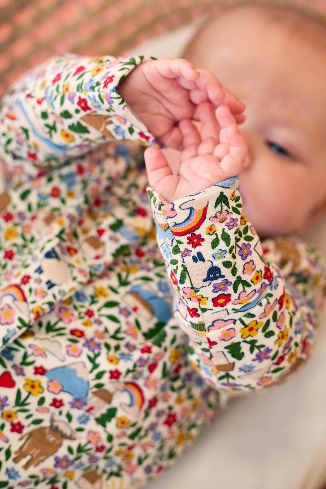 Baby girl wearing a Frugi organic cotton floral babygro with rainbows and mountains.