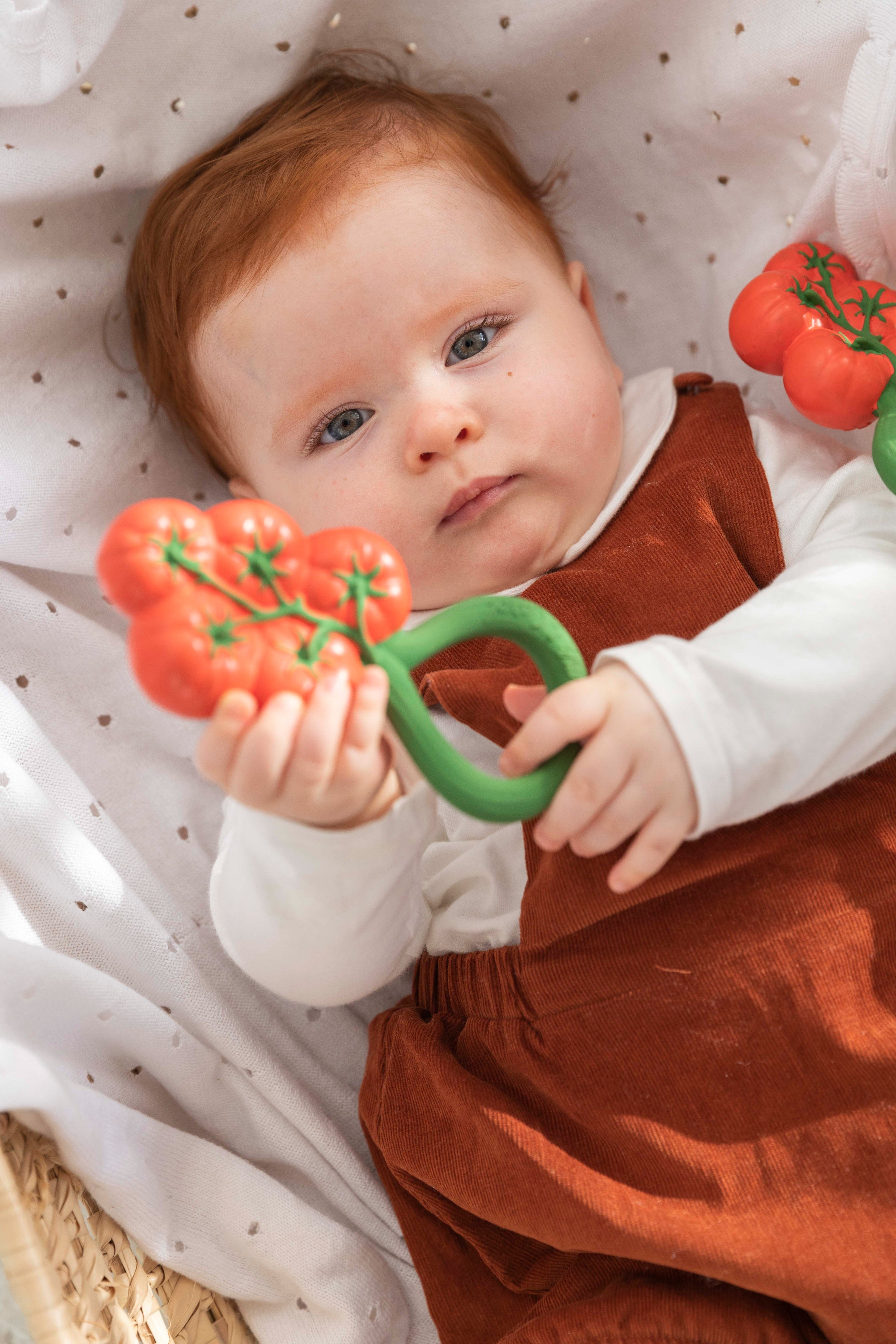 Baby holding a 2-in-1 baby toy, tomato rattle with upcycled cherry seeds, gentle sounds, and natural rubber teether.
