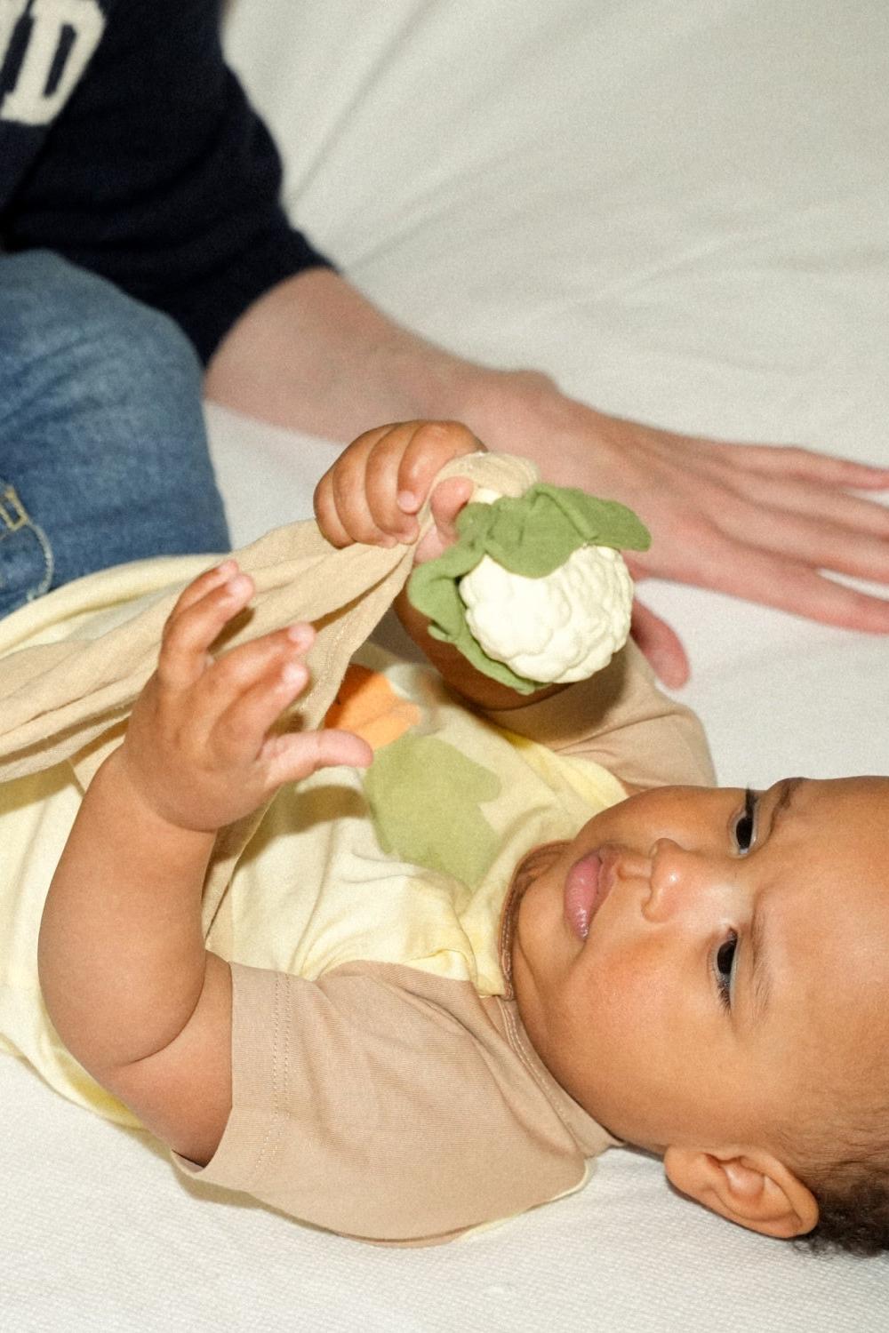 Baby playing with an organic cotton baby comfort toy, Chloe the Cauliflower, with soft leaves and rubber veggie detail.