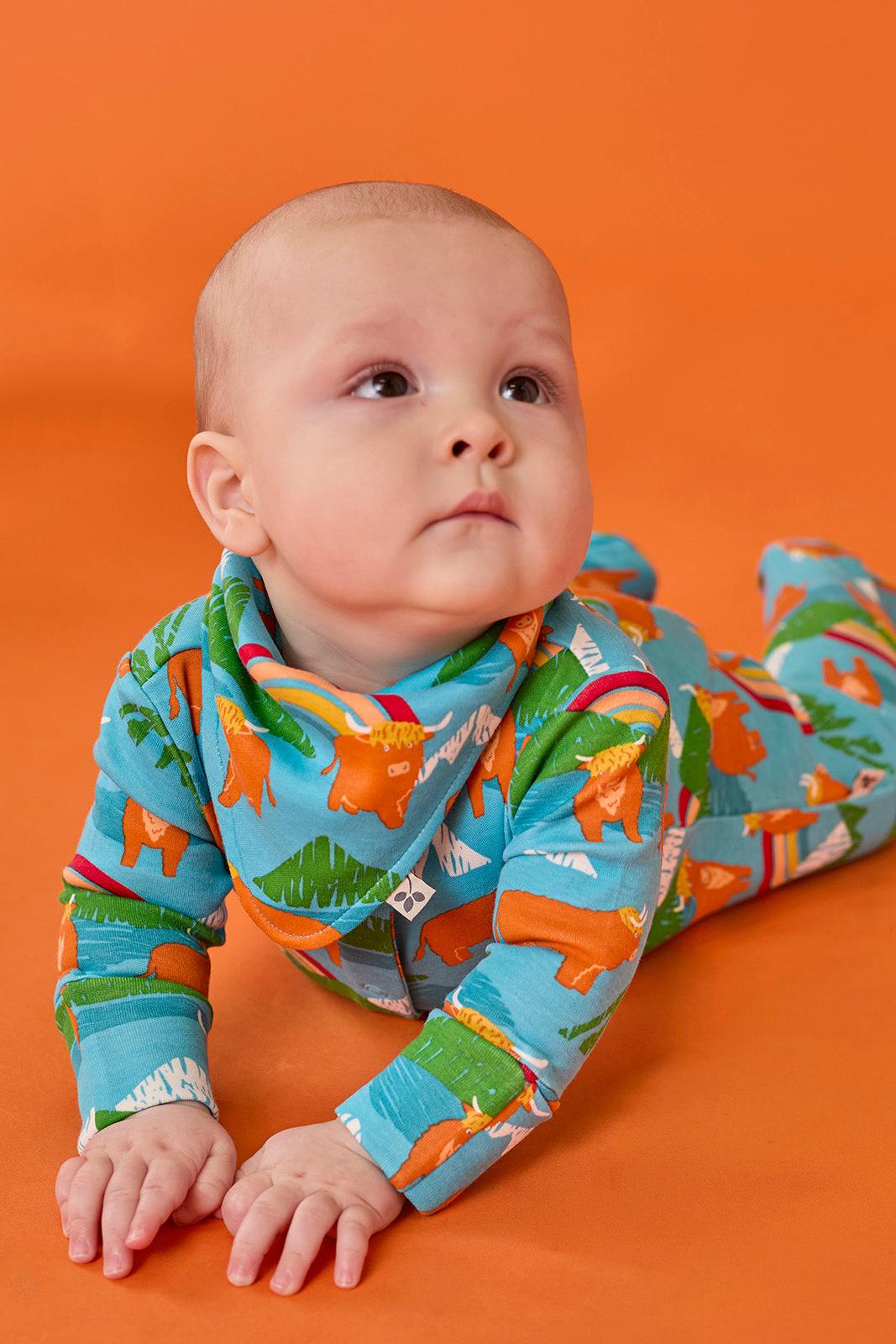 Baby wearing a blue Frugi organic cotton babygro with a cow and farm print, lying on an orange background.