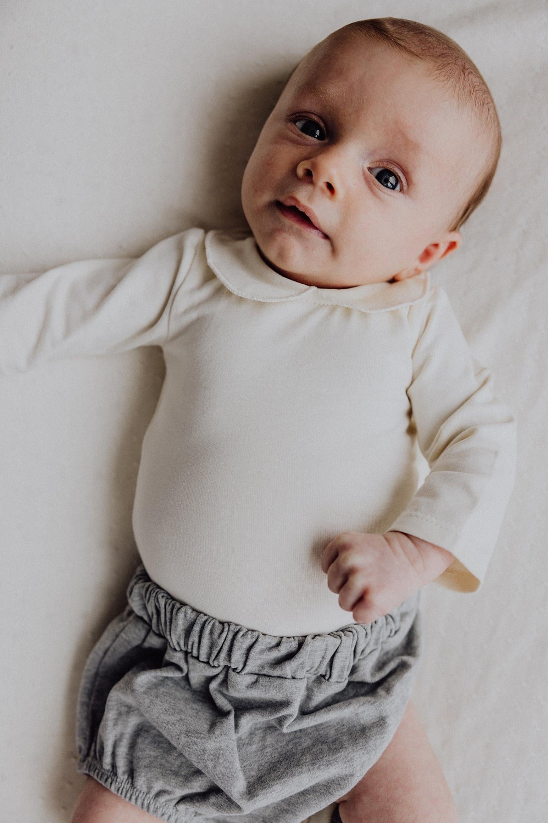 A baby lying on a light cream fabric surface, wearing a cream long-sleeve bodysuit and gray bloomers.