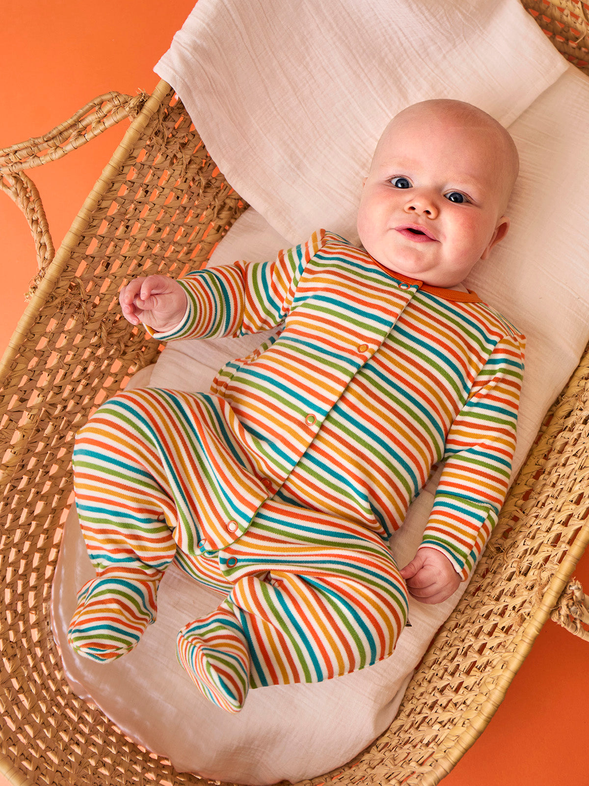 A happy baby wearing a colorful stripe organic cotton babygro lying in a woven wicker moses basket.