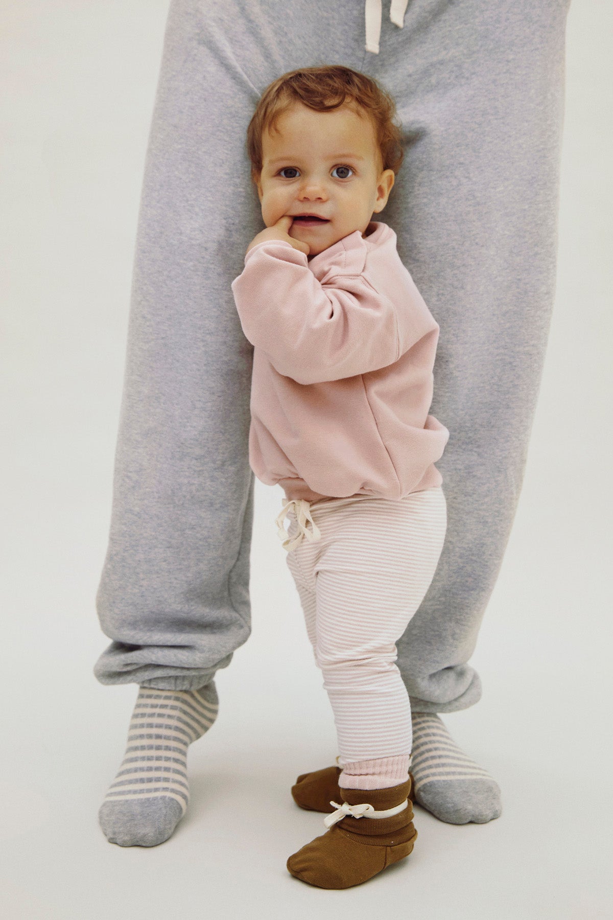 A baby wearing a dusty pink long-sleeve top, white and pink striped pants, and brown booties stands between an adult's legs wearing gray sweatpants and gray striped socks, against a plain white studio background.