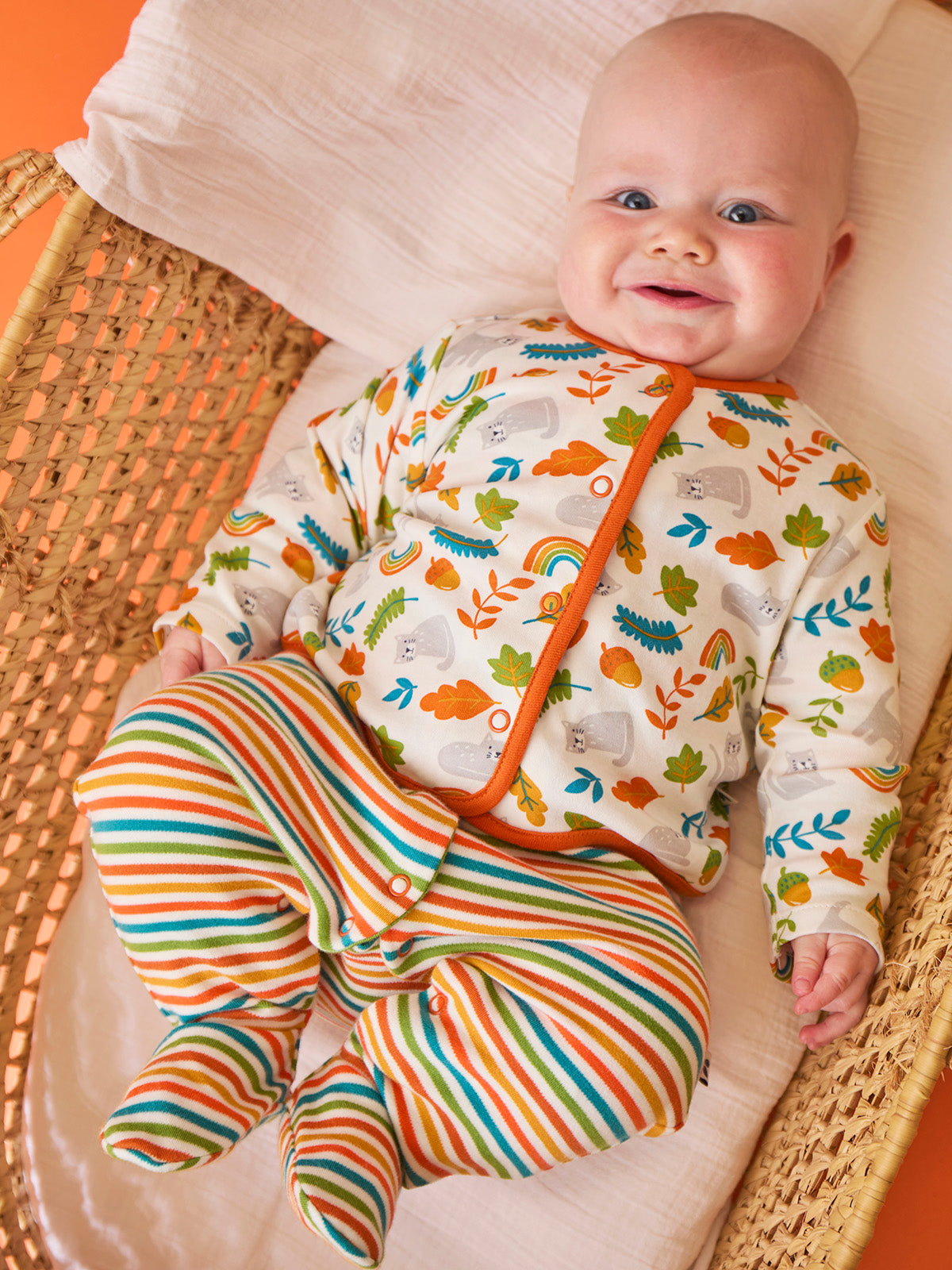 A smiling baby wearing a Frugi organic cotton babygro with autumn leaf and cat print inside a wicker basket.