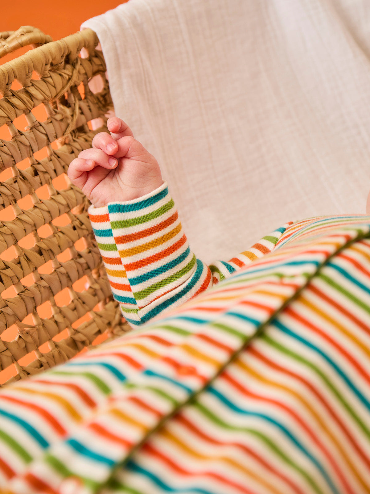A baby hand wearing a Frugi multi-colored stripe organic cotton babygro while resting in a wicker basket.