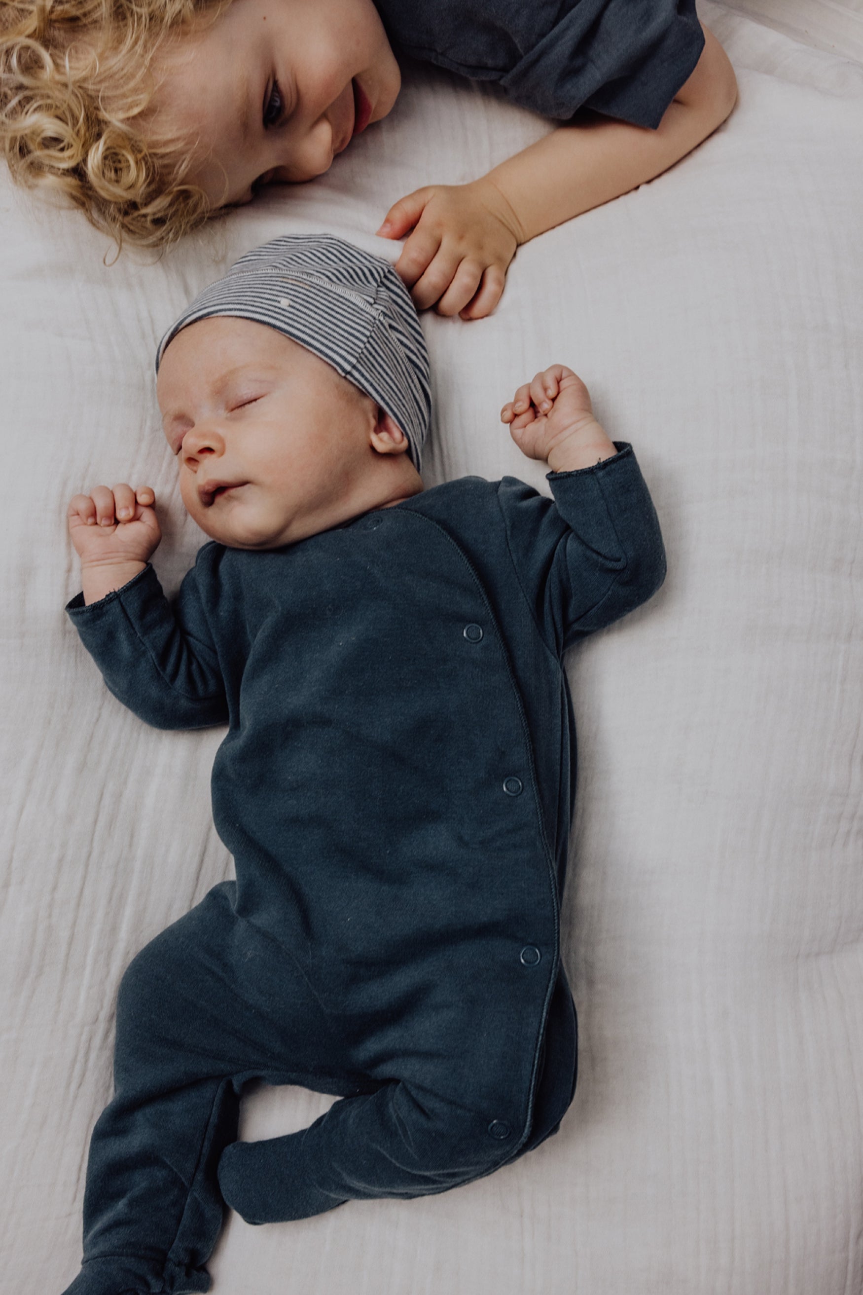 A sleeping newborn baby wearing a dark navy long-sleeve footed romper and a gray striped beanie lies on a white textured blanket, with a toddler in a matching dark navy romper resting above them.