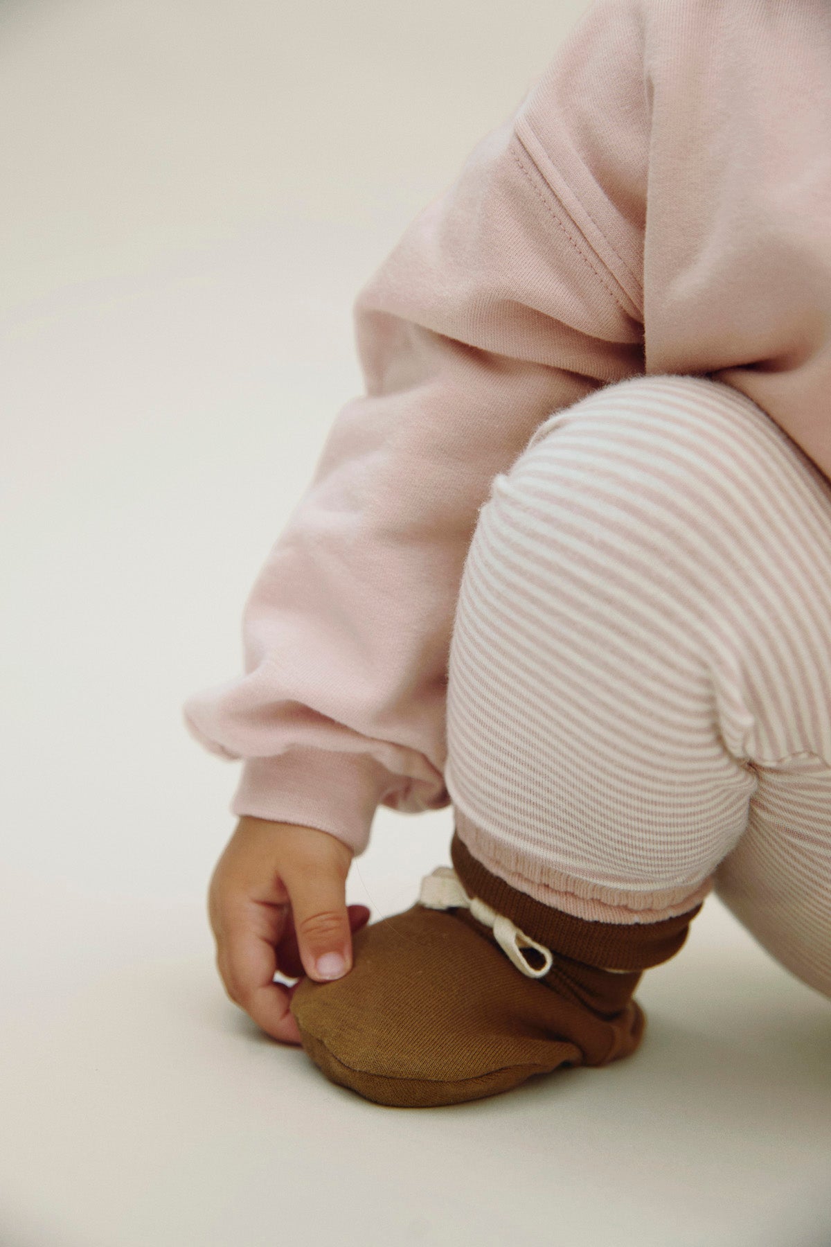 A close-up of a child wearing a pale pink sweatshirt, cream and pink striped pants, and a brown soft bootie, with the child's hand adjusting the bootie against a plain light background.