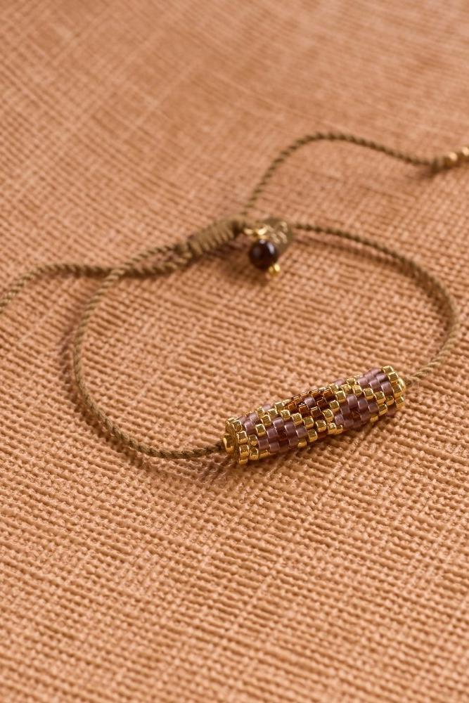 A delicate women's bracelet featuring a woven bead cylinder on an adjustable cord, resting on a textured fabric surface.