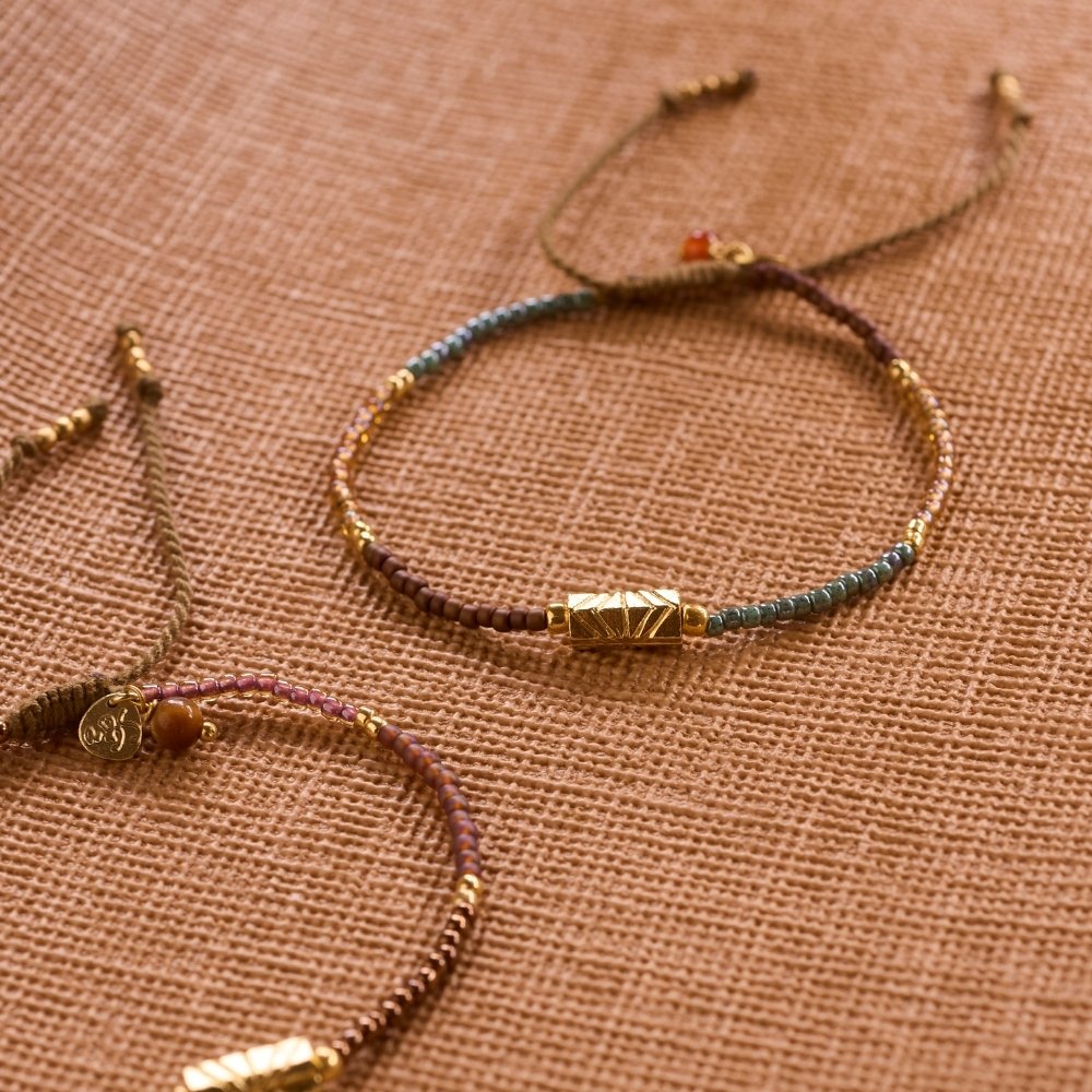 Two handmade beaded bracelets with gold accents and carnelian gemstones on a textured beige fabric background.