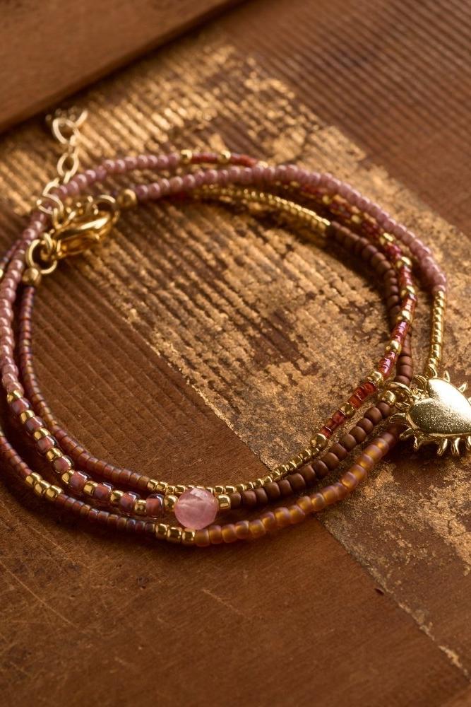 A delicate beaded gold bracelet featuring a small gold heart charm and a soft rose quartz gemstone accent.