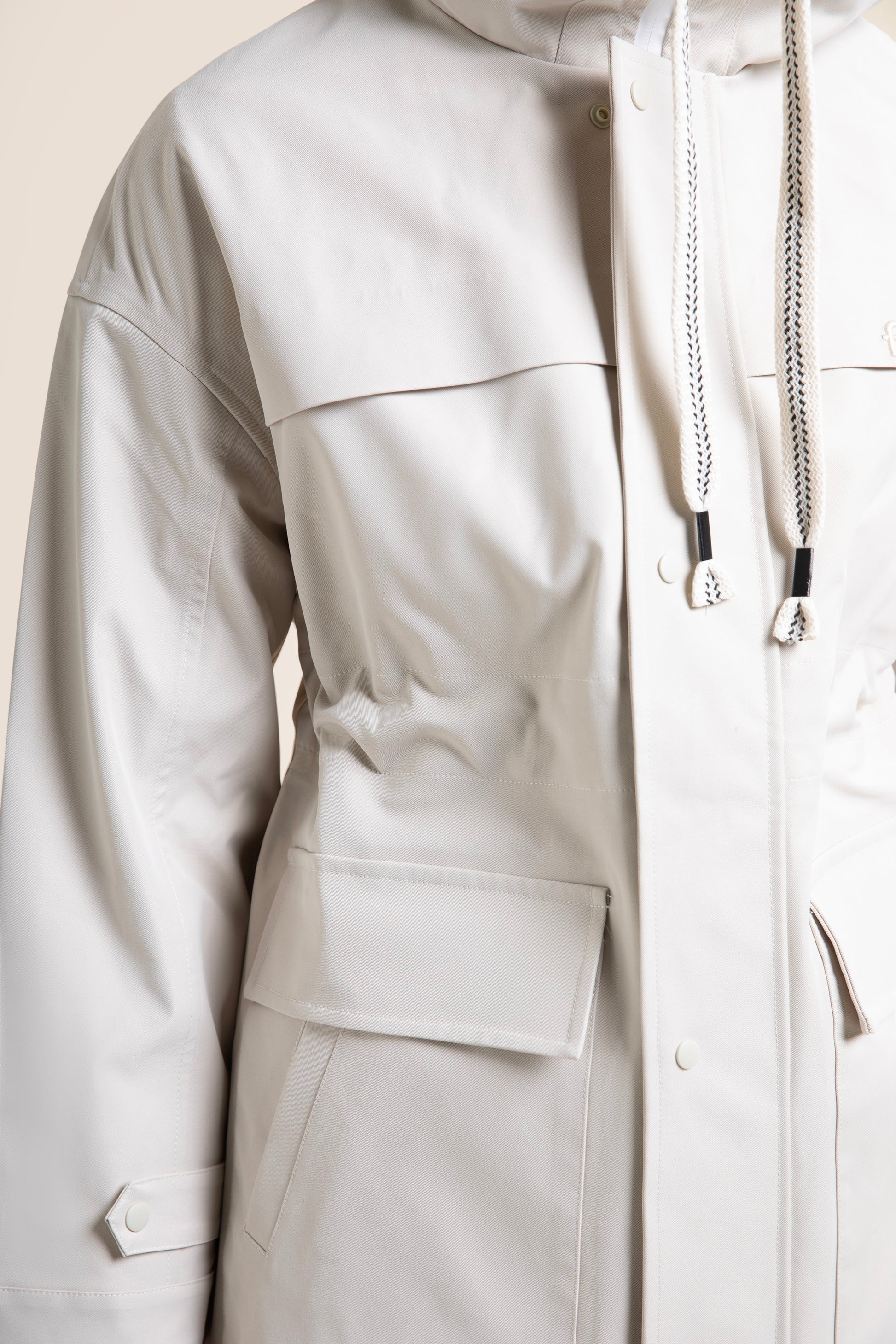 A person wearing a stylish beige Flotte waterproof parka coat with a drawstring hood and large pockets.