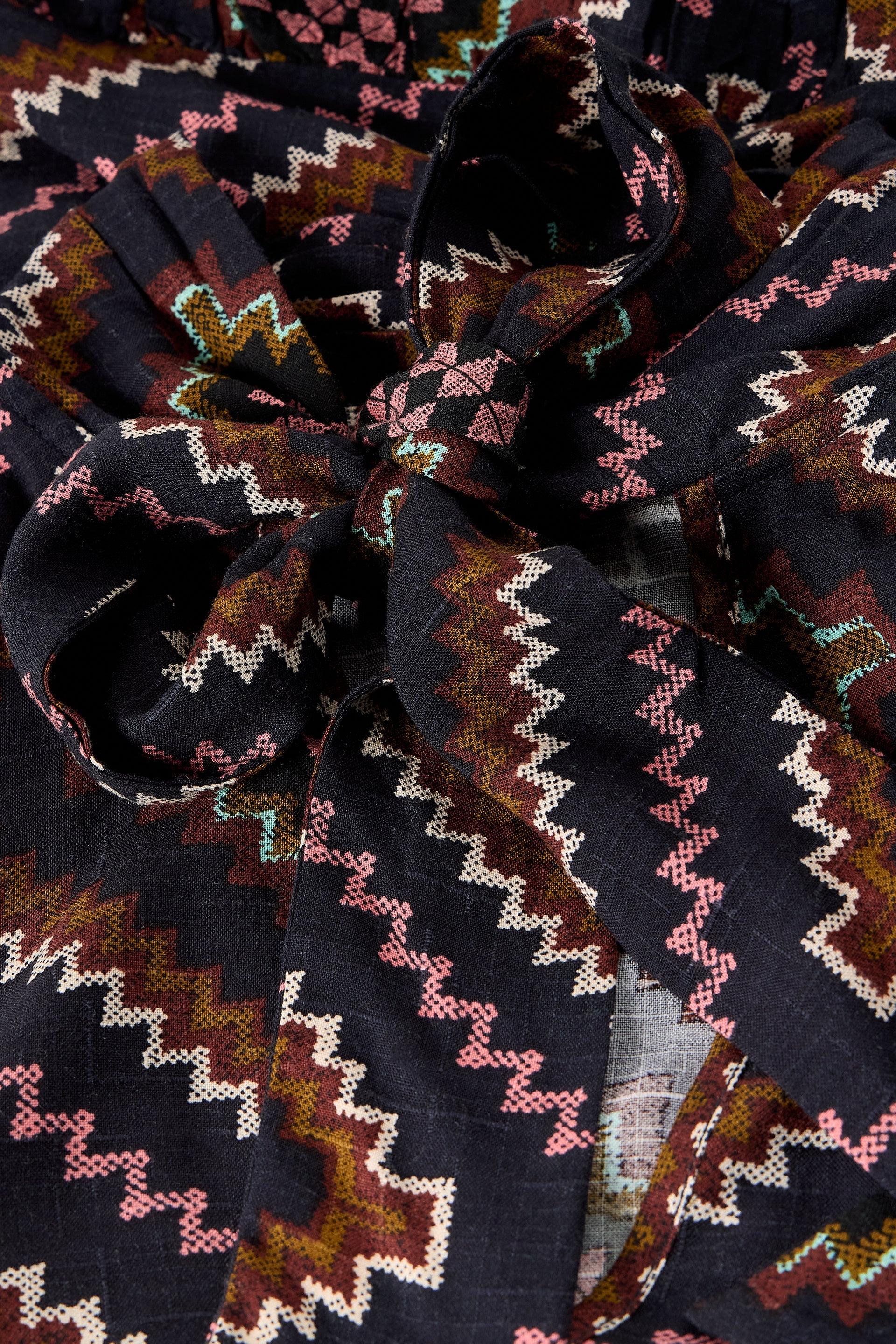 A close-up of a dark woven fabric with a geometric zigzag pattern in brown, pink, cream, and muted green, tied into a bow.