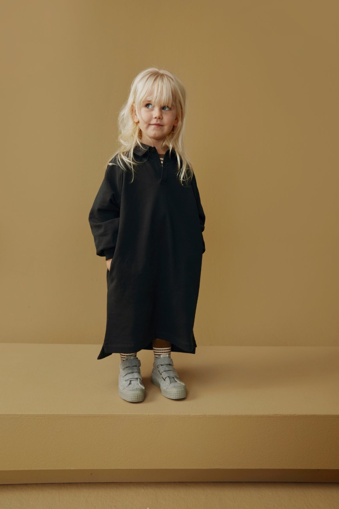 A young child with blonde hair stands on a platform wearing a long black dress with a half-zip neckline and light gray sneakers, set against a solid warm tan background.