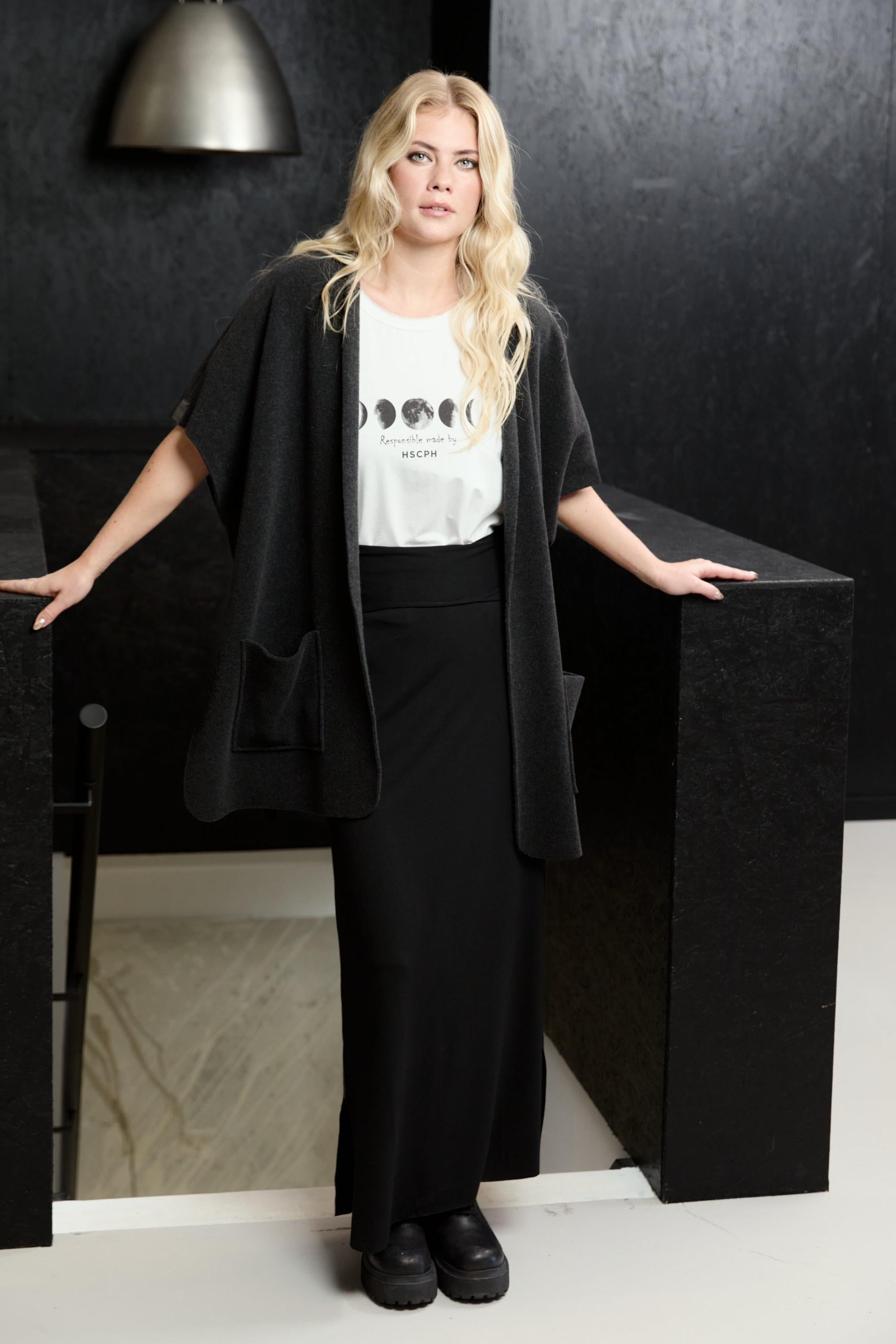 A blonde woman models a black open-front cardigan over a white graphic T-shirt and black wide-leg pants, standing between black display pedestals in a dark, modern interior.