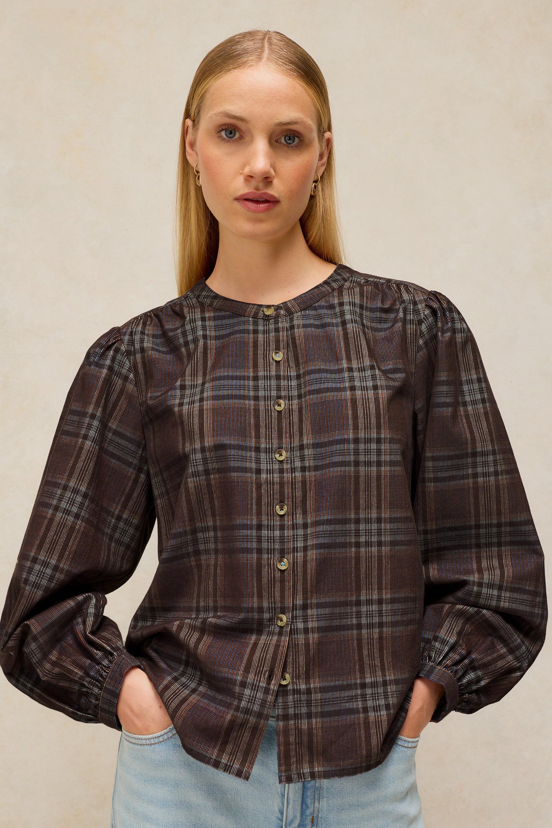 A woman modeling a dark brown and black plaid long-sleeve blouse with gold buttons, paired with light blue jeans, against a neutral beige background.