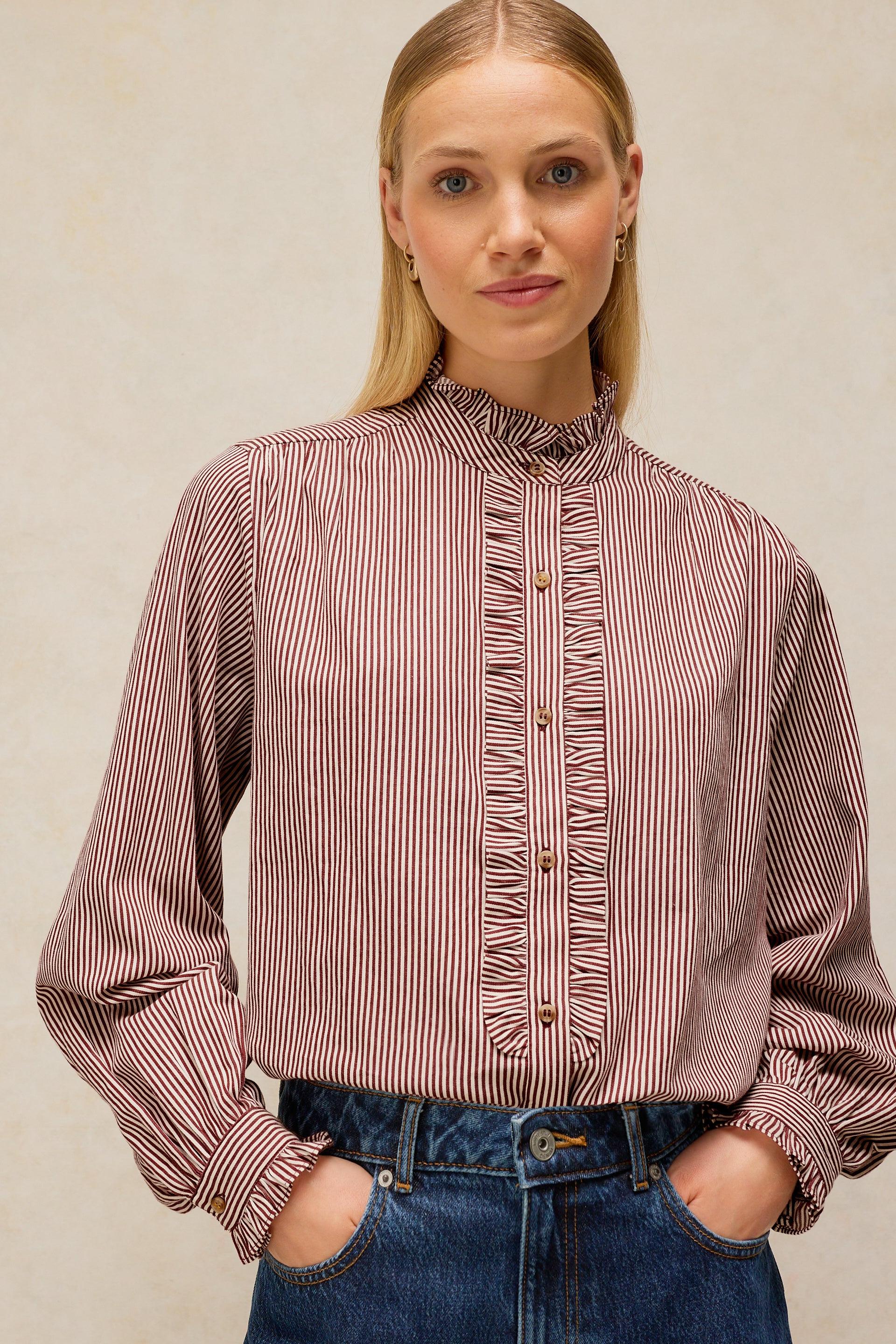 A woman wearing a long-sleeve button-front blouse with vertical stripes in dusty rose and white, paired with blue jeans, against a neutral beige background.