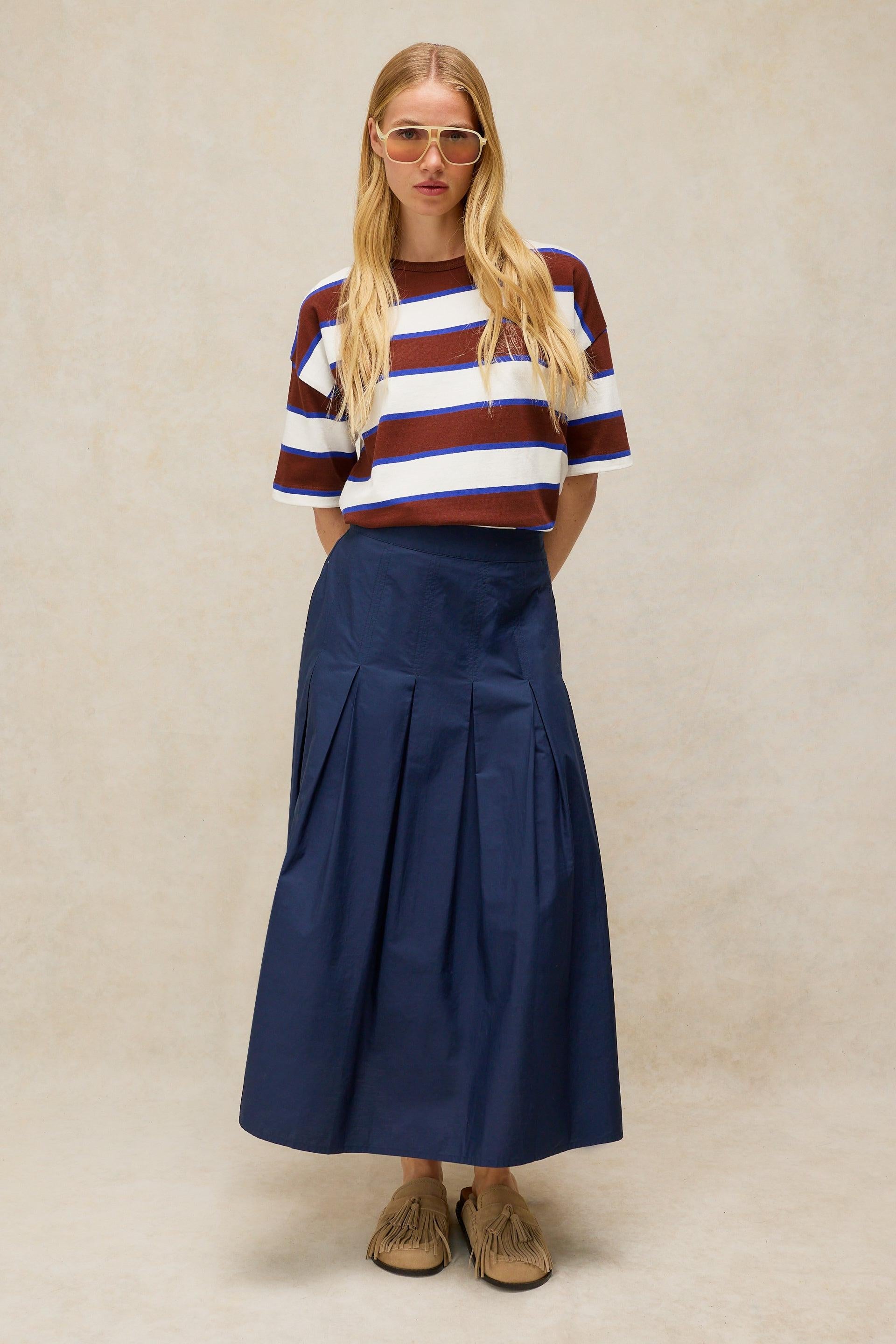 A blonde woman wears a brown and blue striped organic cotton t-shirt and a long navy pleated maxi skirt.