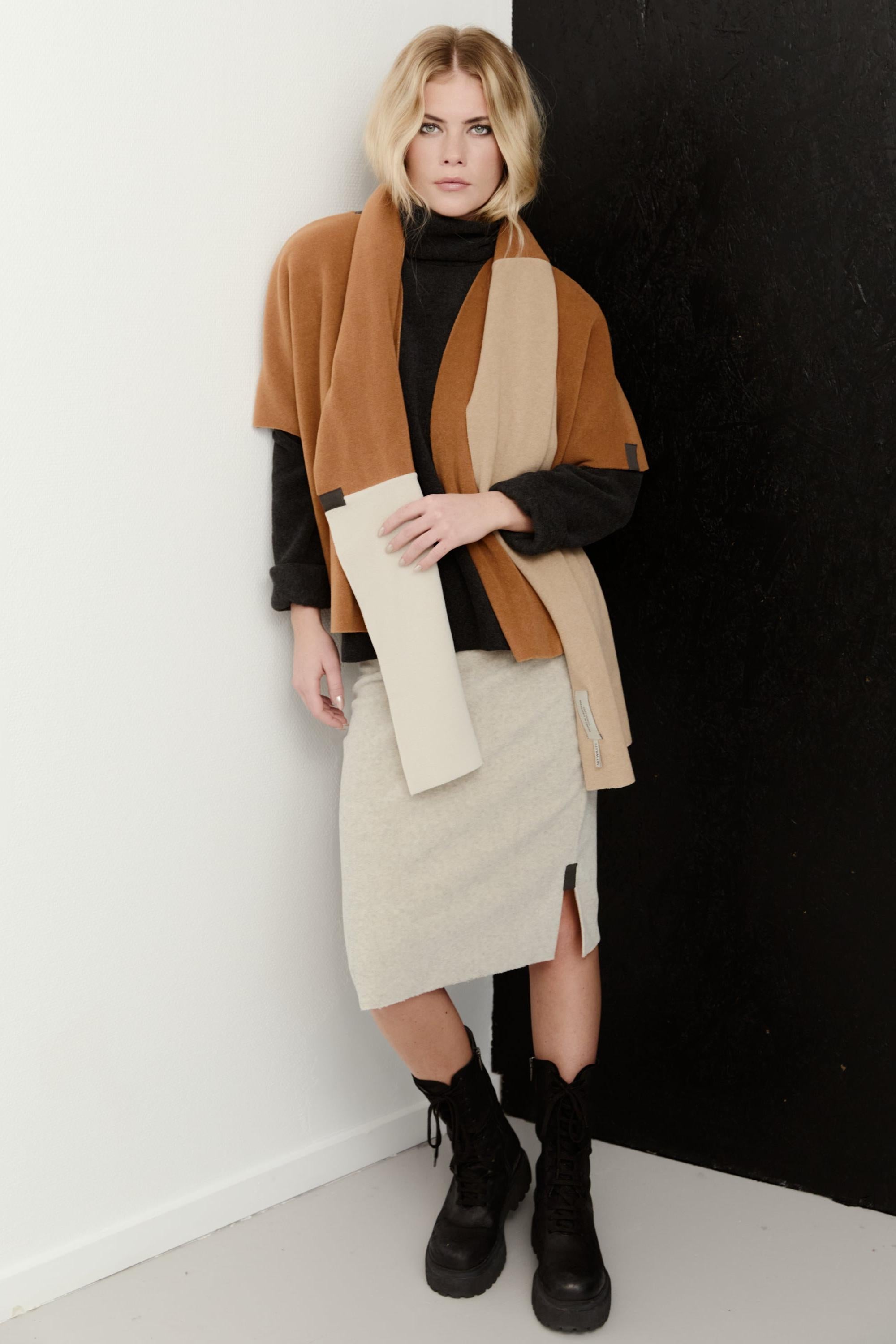 A woman models a stylish outfit consisting of a brown and beige color-blocked wrap jacket, a black turtleneck, a light beige midi skirt, and black lace-up ankle boots, standing against a split white and black wall.