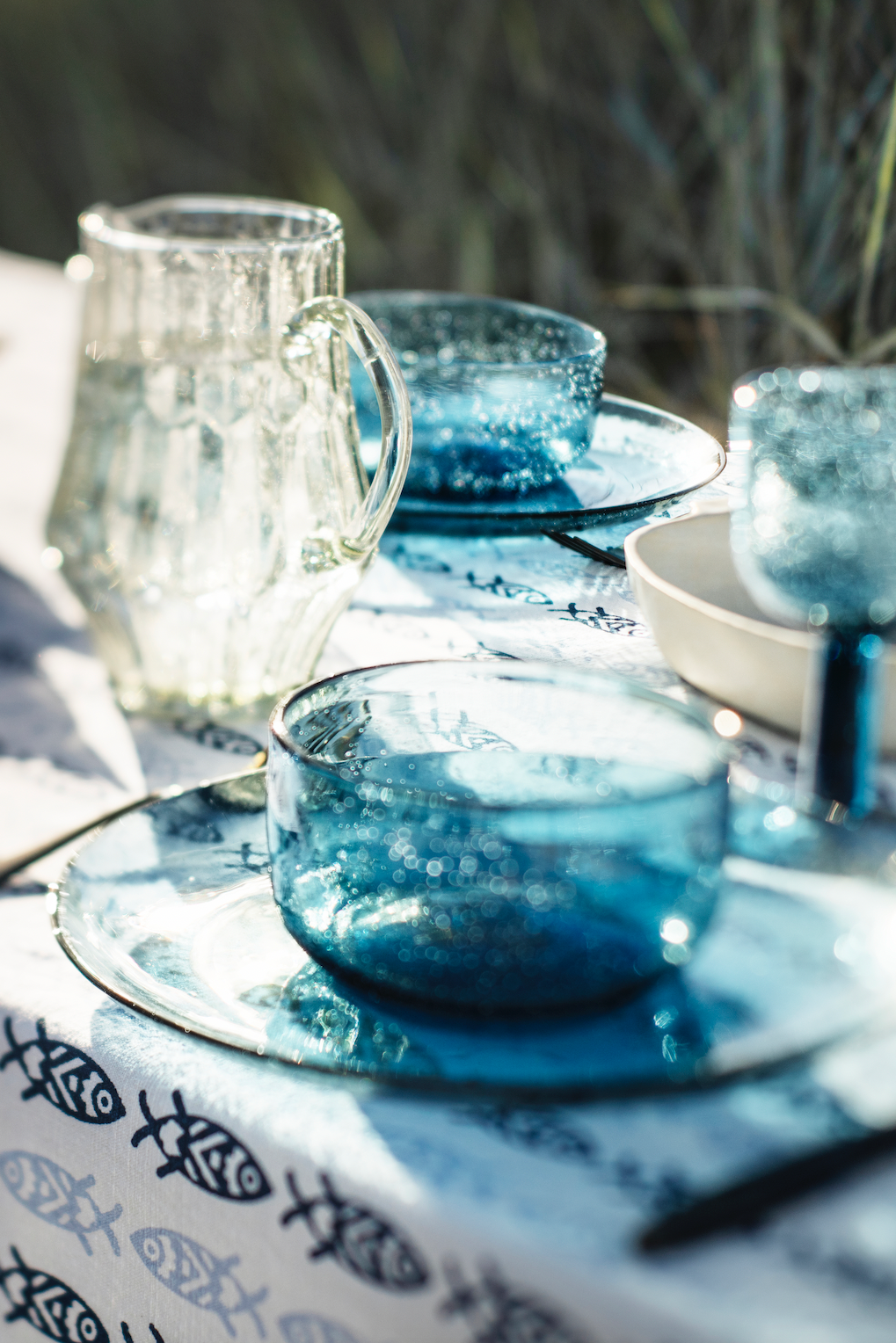 Blue glass bowls and pitcher on a white cotton tablecloth with a fish pattern.