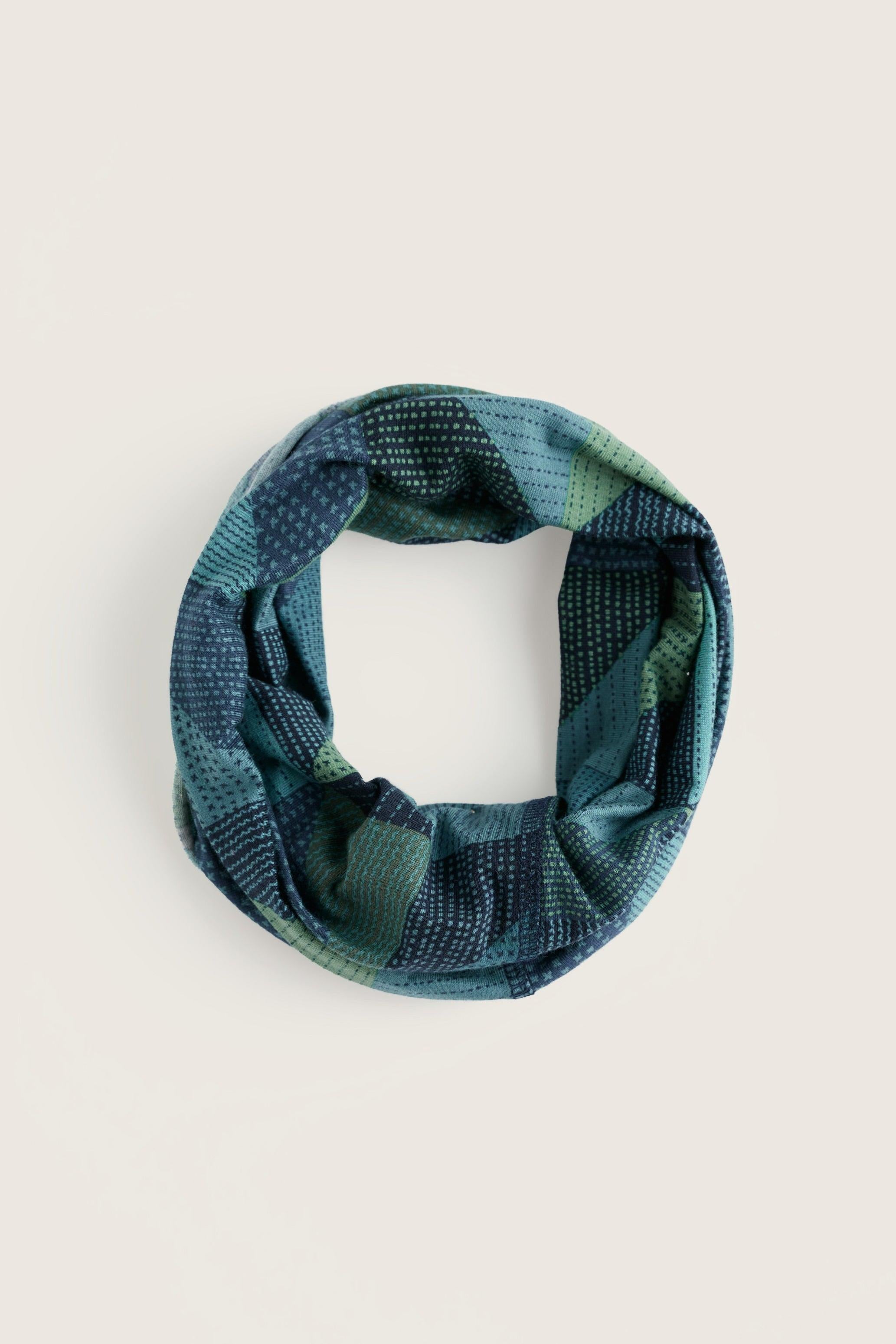 A patterned infinity scarf in shades of teal, blue, and green, displayed on a solid light beige background.