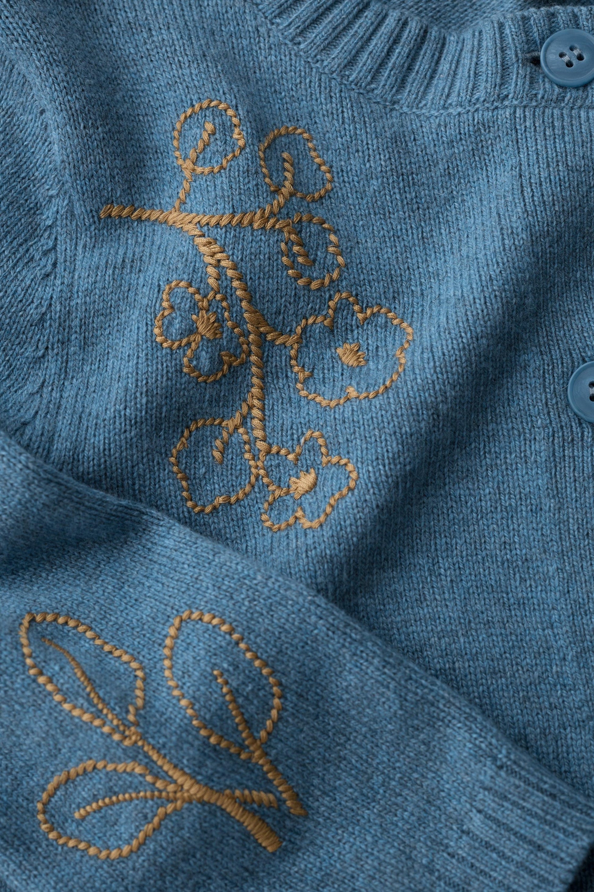 Close-up of a blue knit cardigan sweater with gold embroidered floral and leaf motifs and blue buttons.