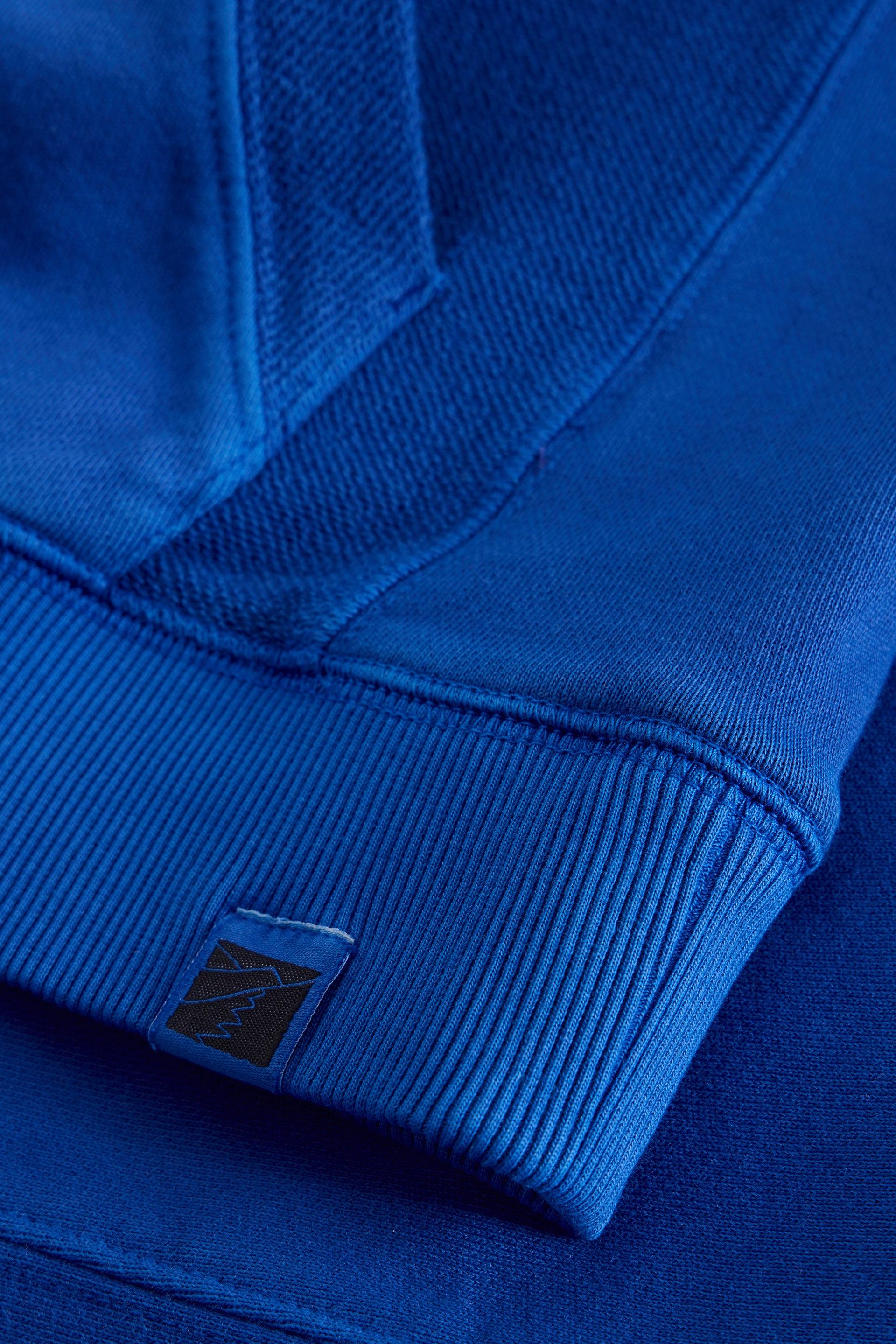 Close-up of a royal blue sweatshirt cuff with a small square brand patch on the ribbed hem.