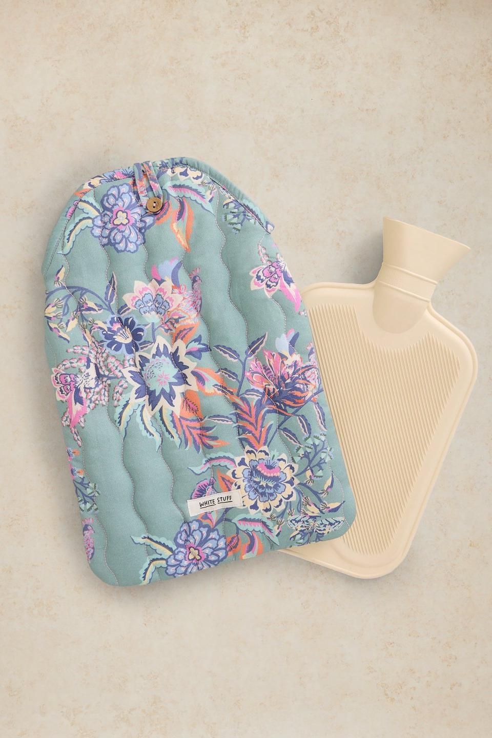 A quilted hot water bottle cover in a floral print with a small button closure, shown next to a cream-colored hot water bottle on a textured beige background.