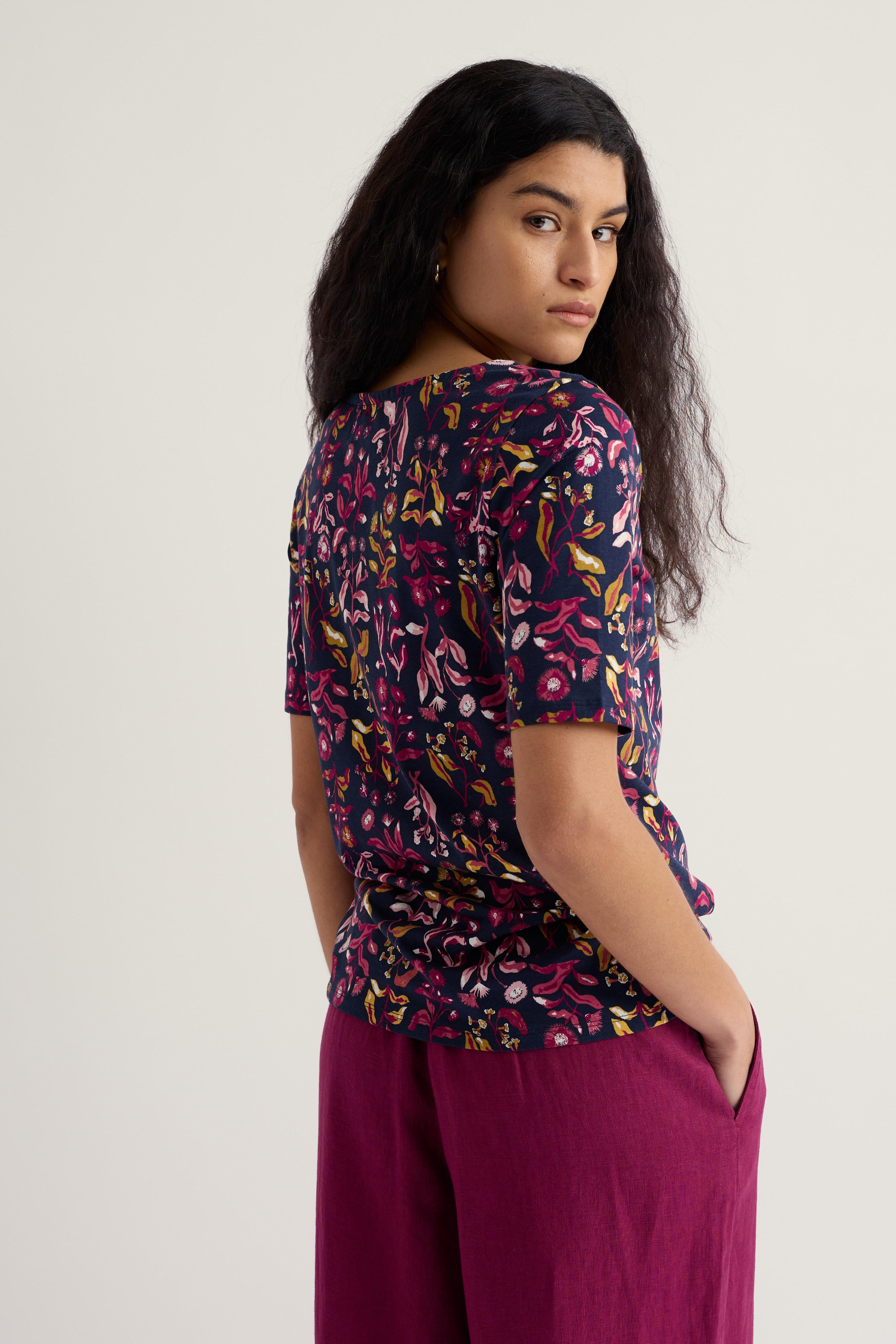 Woman wearing a patterned organic cotton top and purple pants against a plain background