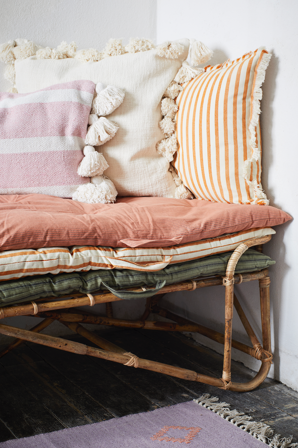 Rattan bench with layered cushions, decorative pillows, and a lilac area rug in a minimalist room.