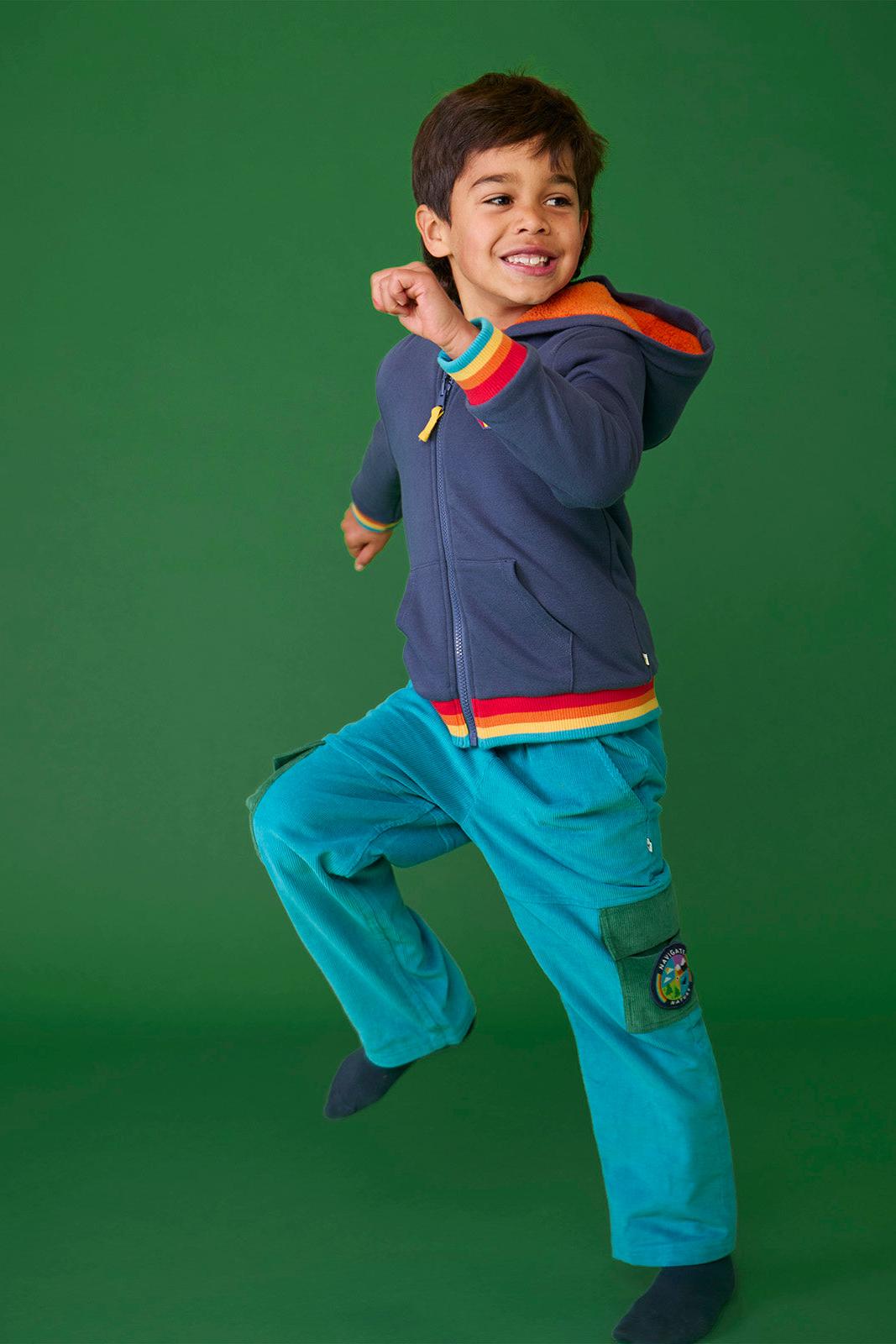 A happy young boy wearing teal Frugi organic cotton cord trousers jumping against a green background.