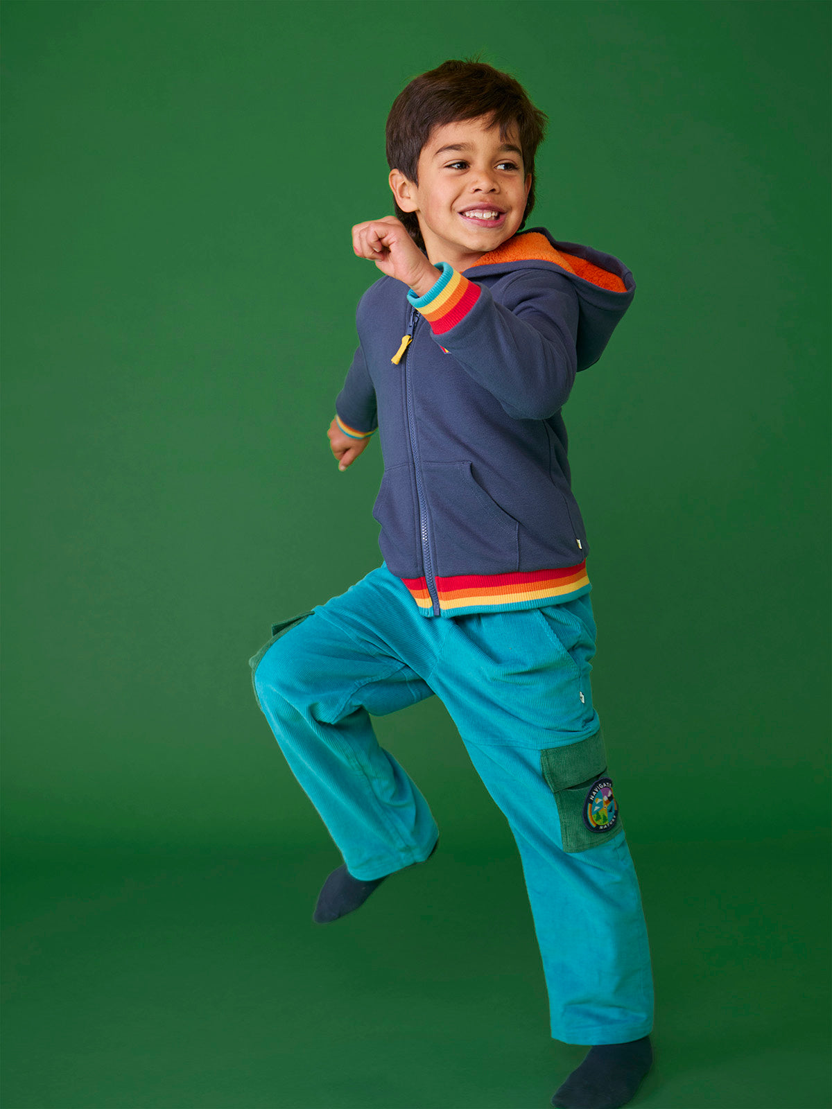 A happy young boy wearing teal Frugi organic cotton cord trousers jumping against a green background.