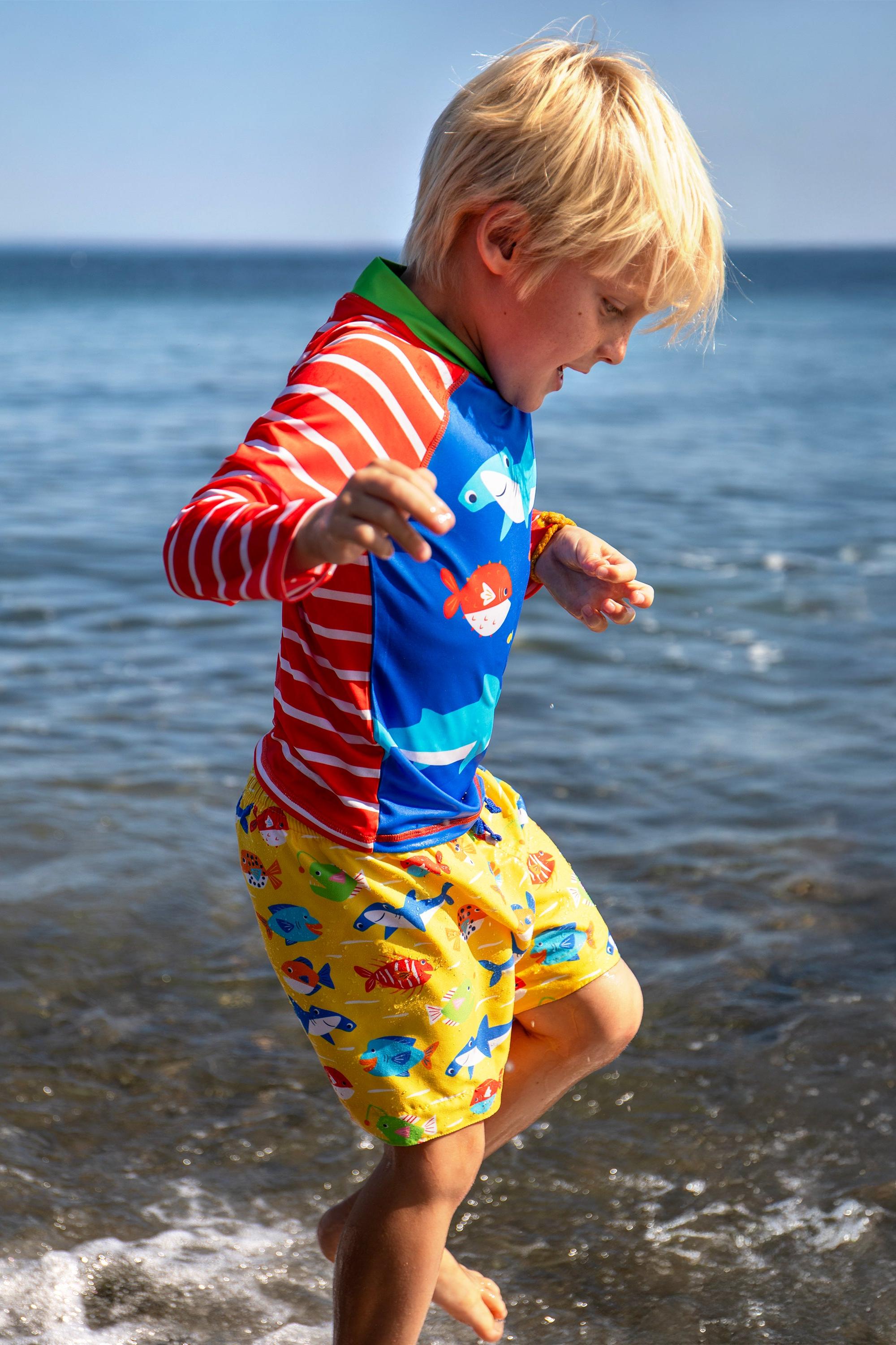A young boy with blonde hair wearing colorful Frugi fish-patterned swimwear playing in the sea.