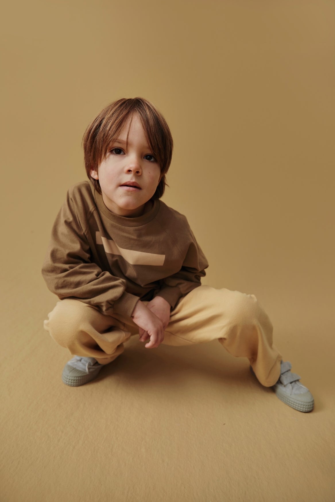 A young child crouching in a studio setting, wearing a brown sweatshirt with a beige chest stripe, beige pants, and light blue sneakers against a solid tan backdrop.